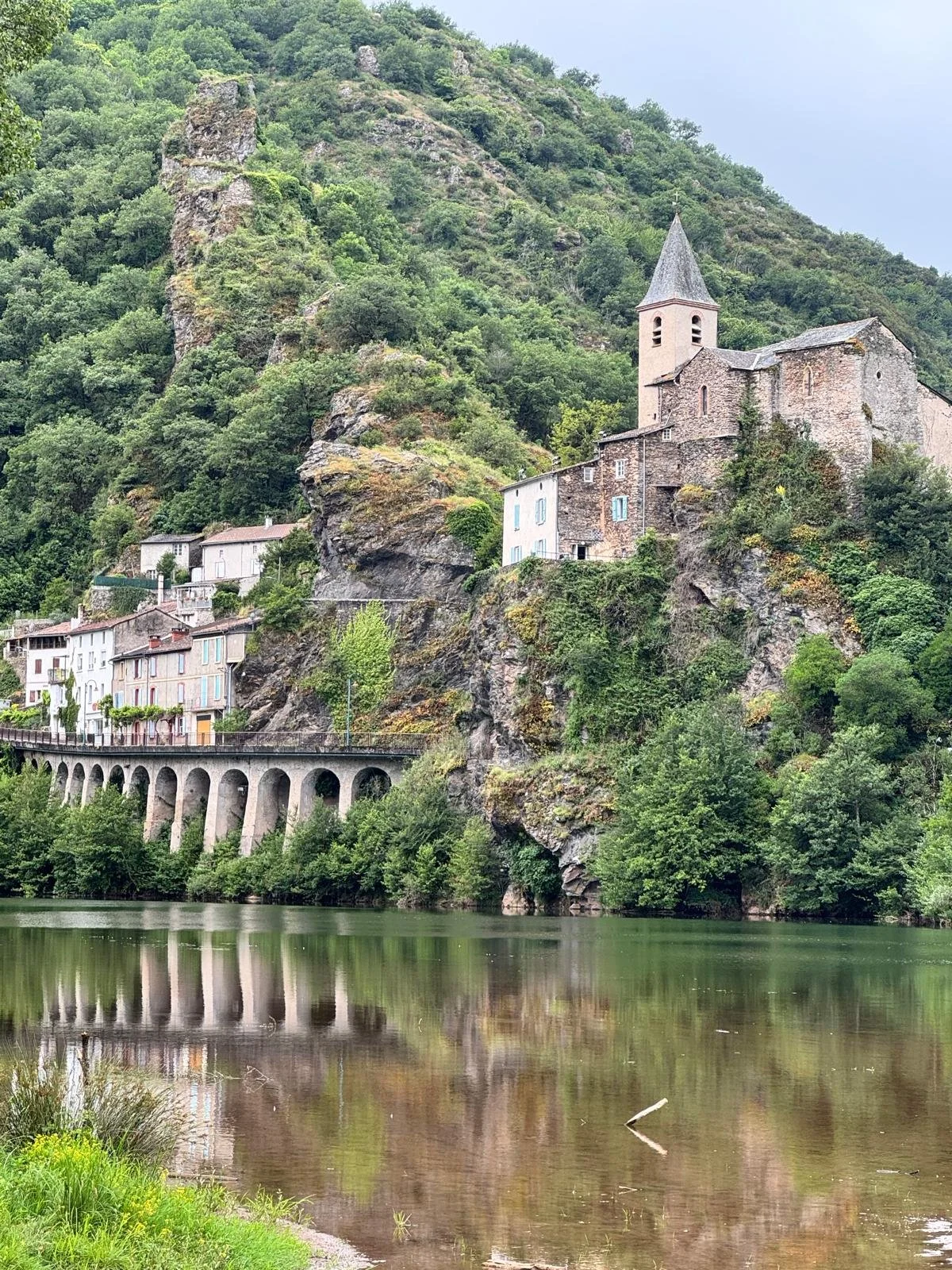 Village perché sur une falaise avec une église, au-dessus d'une rivière calme, entouré de forêt verte.