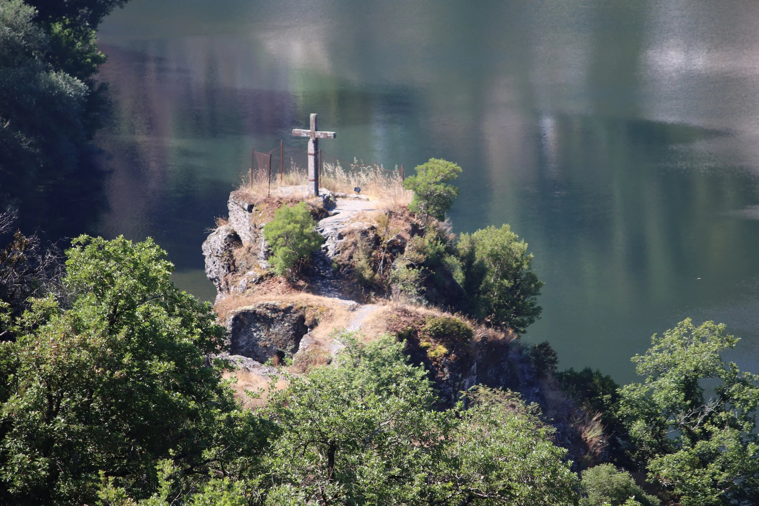 Un rocher au milieu d'un lac avec une croix en bois au sommet, entouré d'arbres verts.