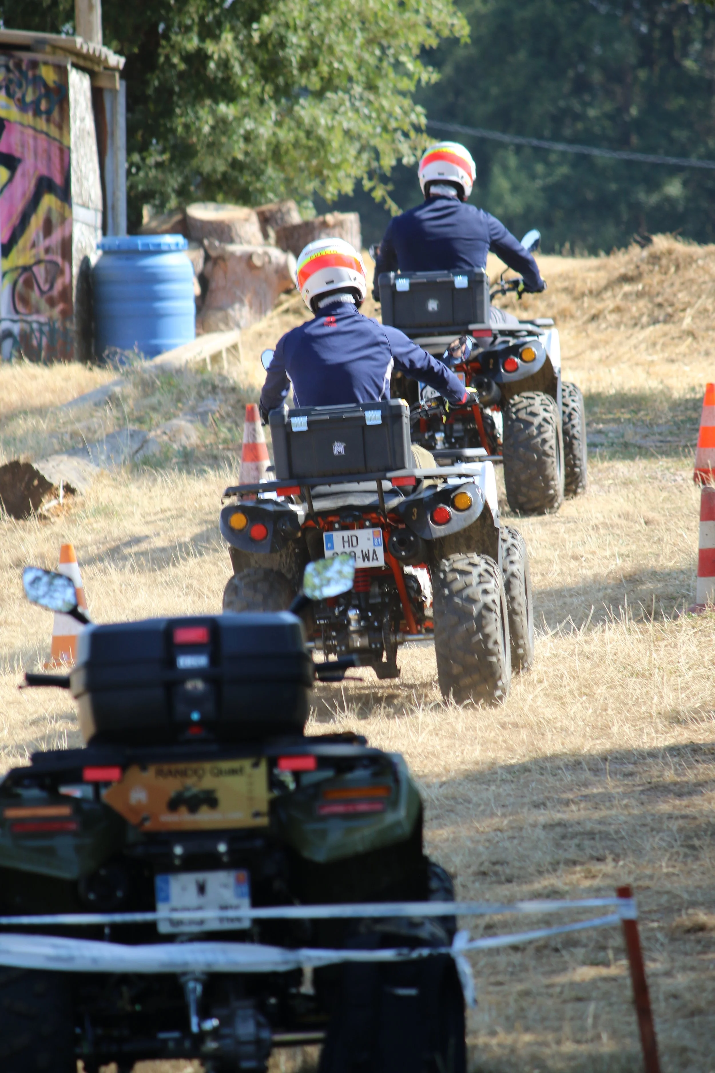 Deux personnes en casques conduisent des quads sur un terrain poussiéreux avec des cônes de signalisation orange, arbres en arrière-plan et un bâtiment avec des graffitis.