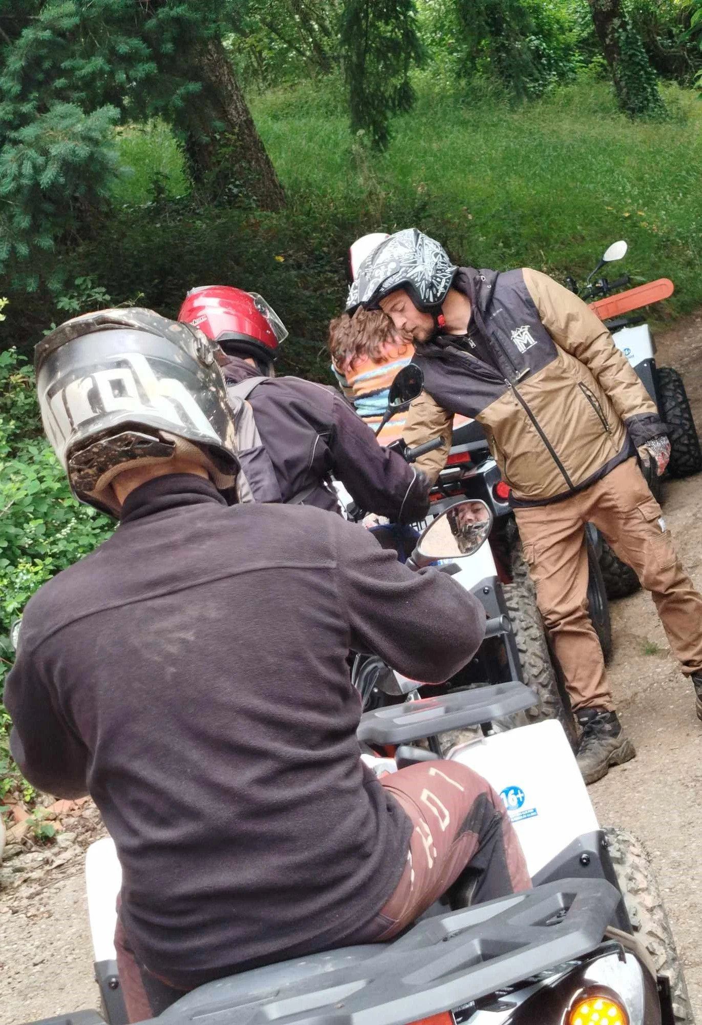 Trois personnes avec des casques de moto, deux debout et une assise, discutant dans une forêt.