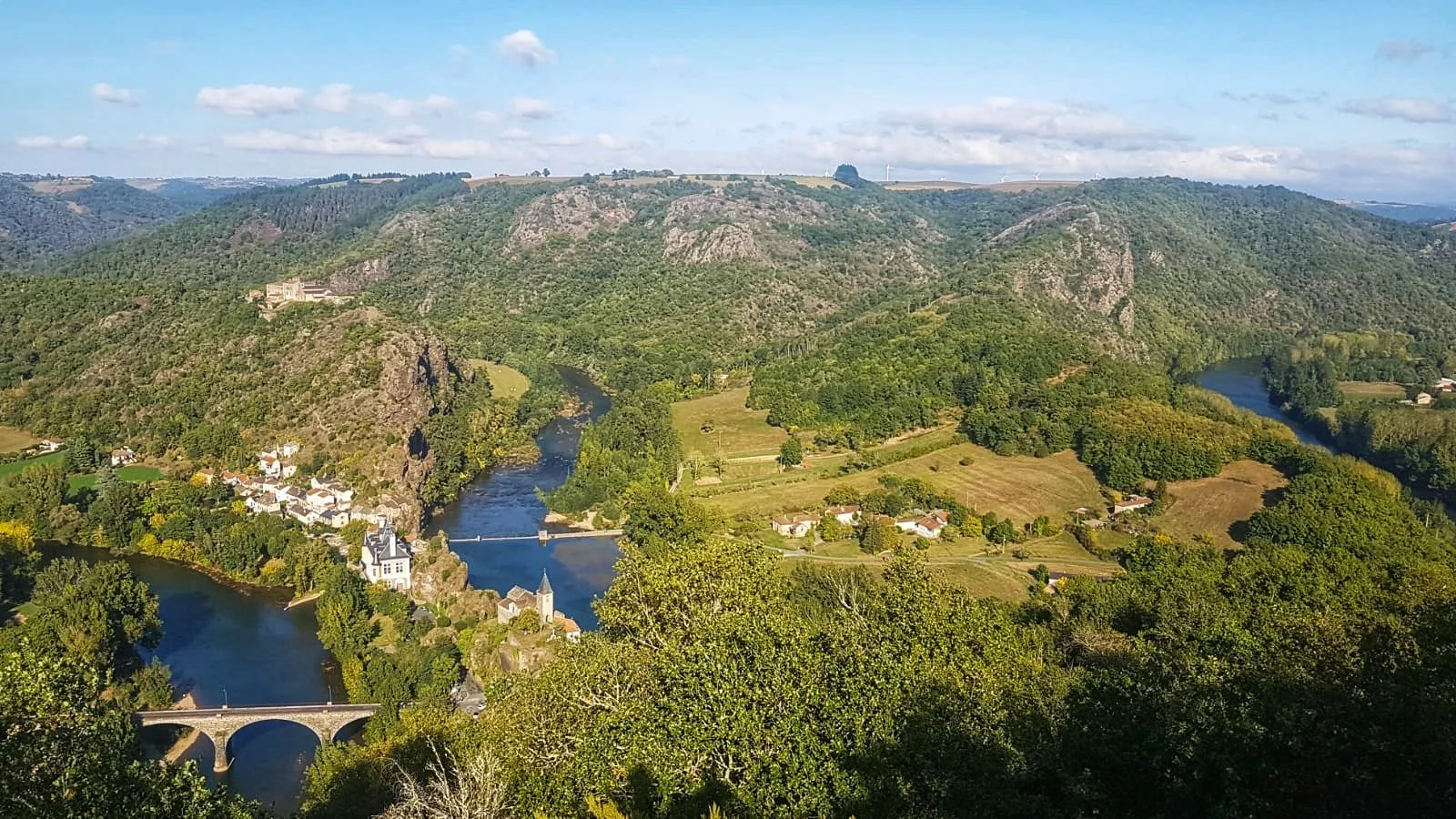 Paysage de vallée verdoyante avec rivières, ponts, maisons, et montagnes sous un ciel partiellement nuageux.