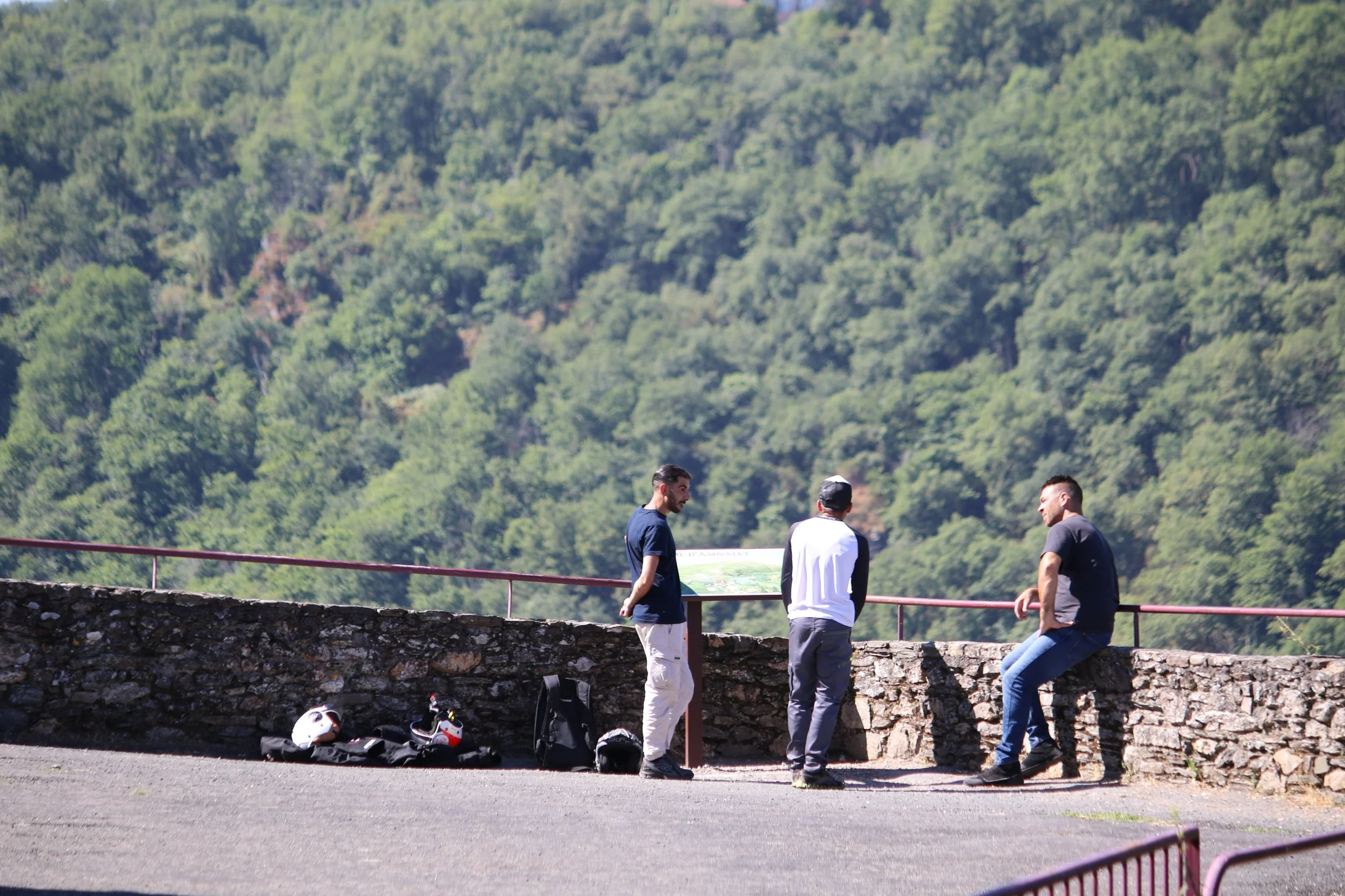 Trois hommes se tiennent devant une vue panoramique de la forêt verdoyante, discutant près d'une barrière en pierre. Deux casques de moto et un sac à dos sont posés au sol à côté d'eux.