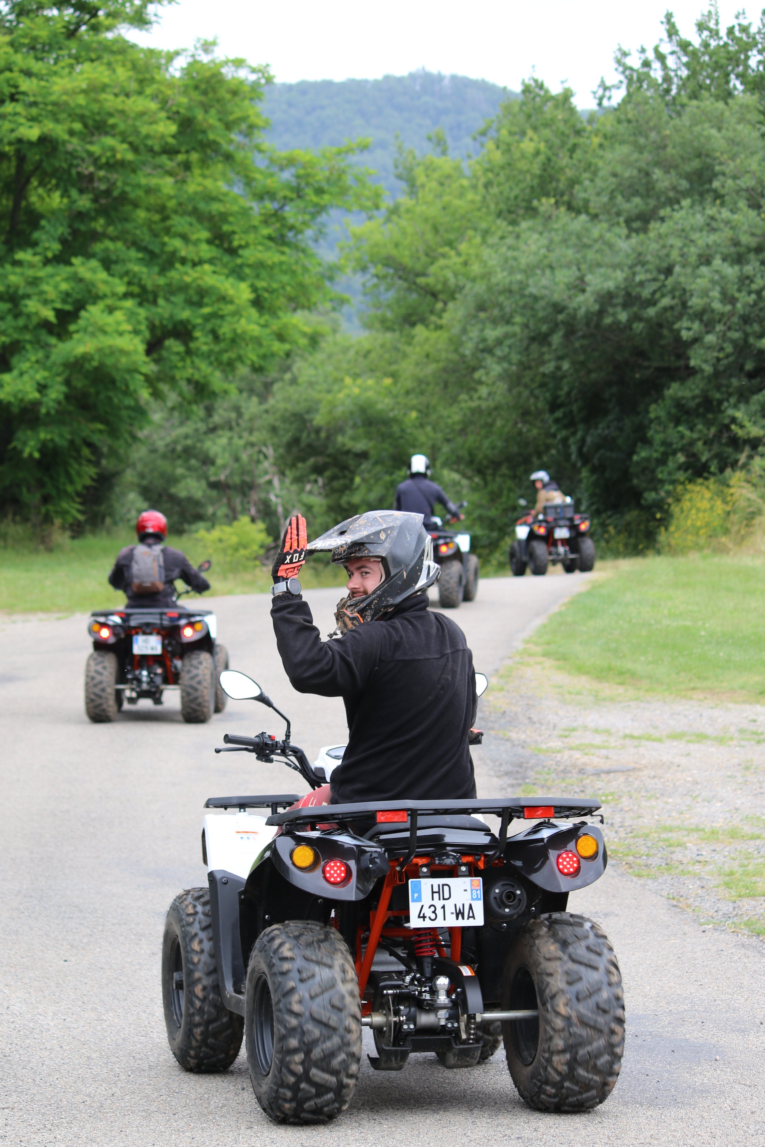 Groupe de personnes en moto quad dans un environnement naturel verdoyant, la personne au premier plan faisant un signe de la main et portant un casque noir.