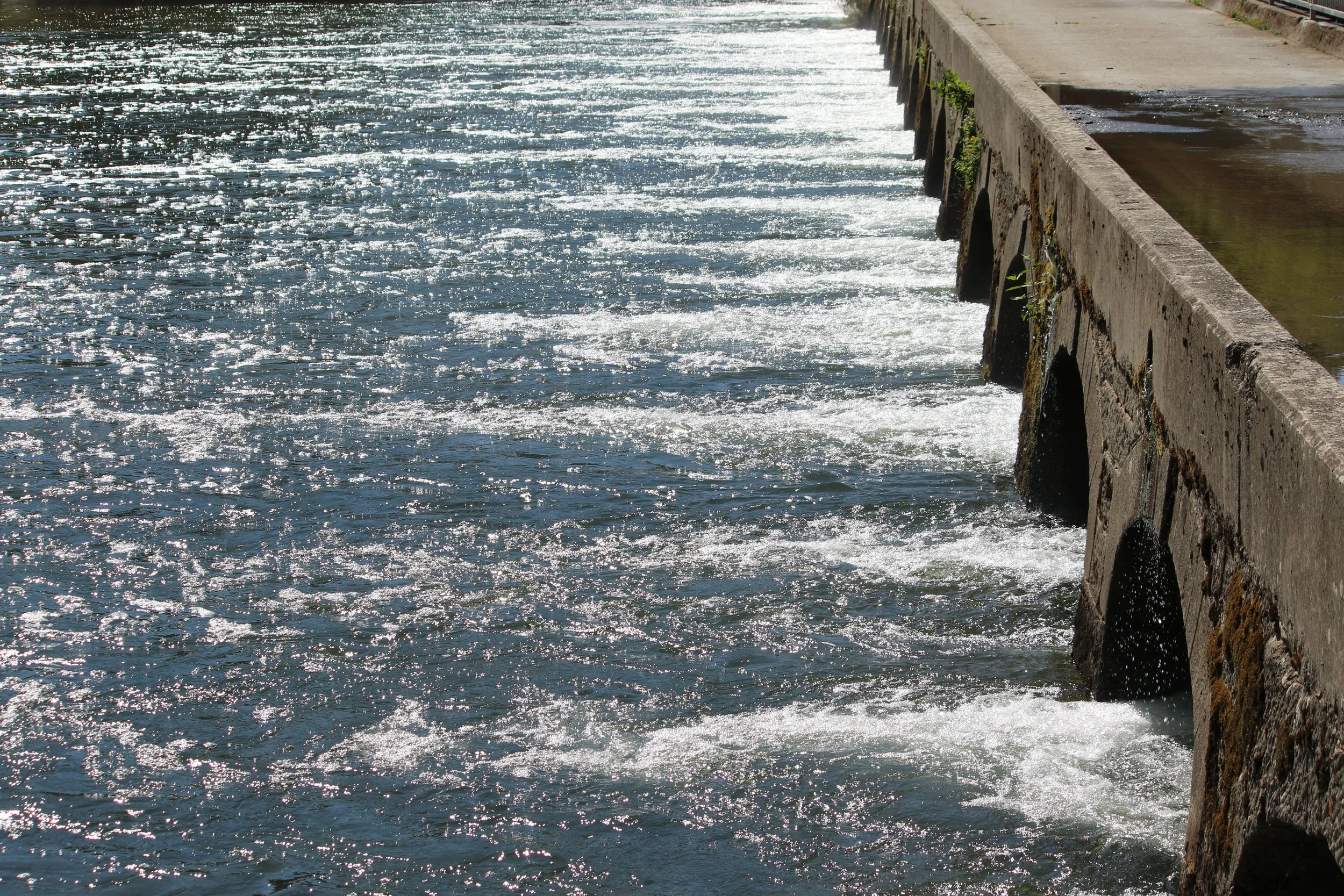 L'eau de la rivière qui coule doucement contre la digue en pierre sur le bord de l'eau, avec des reflets de lumière sur la surface de l'eau.