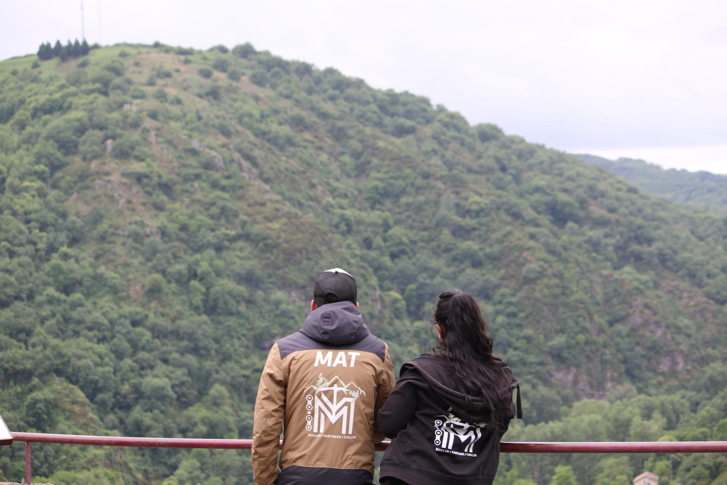 Deux personnes regardant une montagne verte depuis une plateforme en plein air, l'une porte une veste marron et noire avec le mot 'MAT' à l'arrière, l'autre une veste noire avec un logo.