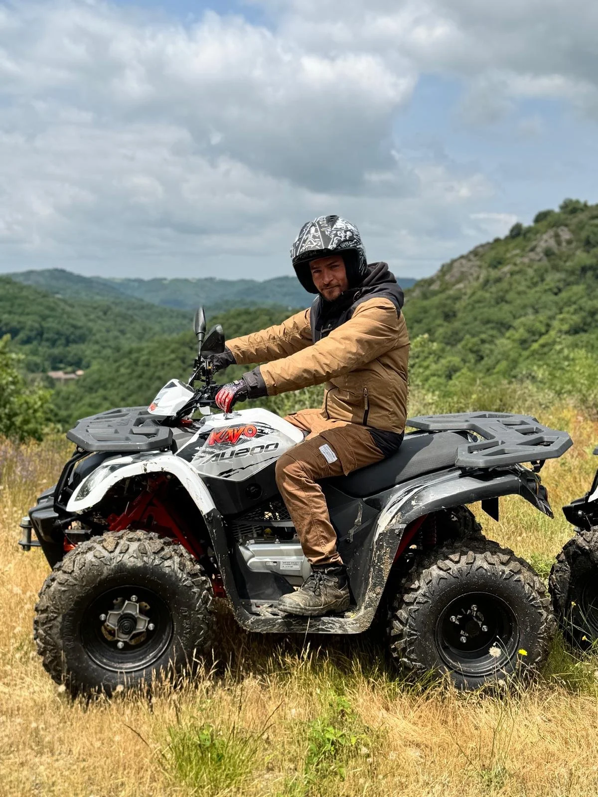 Homme en casque et vêtements de moto tout-terrain assis sur un quad dans un paysage de collines verdoyantes sous un ciel nuageux.