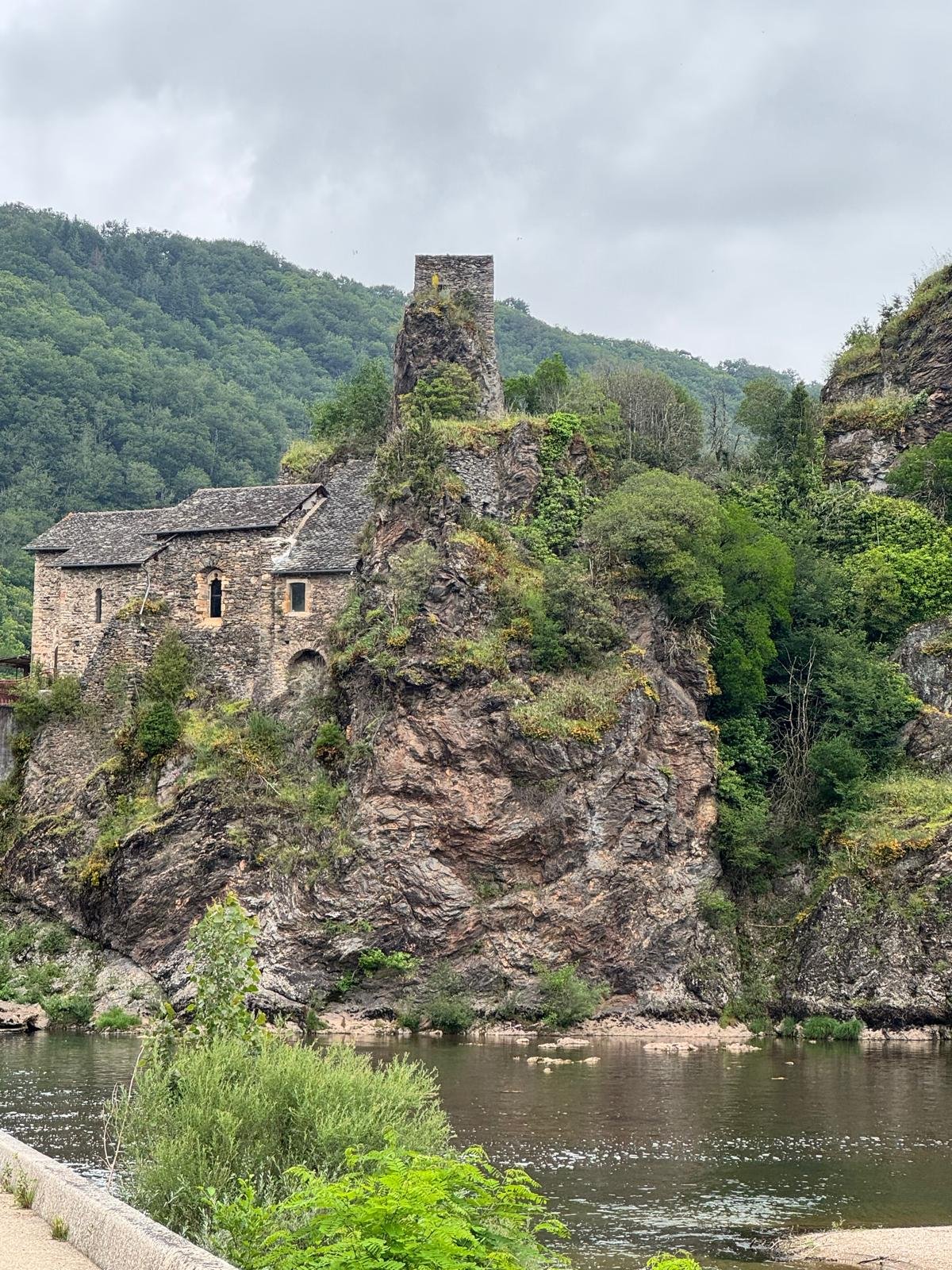 Une vieille abbaye construite en pierre est perchée sur un rocher surplombant une rivière, entourée de collines verdoyantes et de montagnes sous un ciel nuageux.