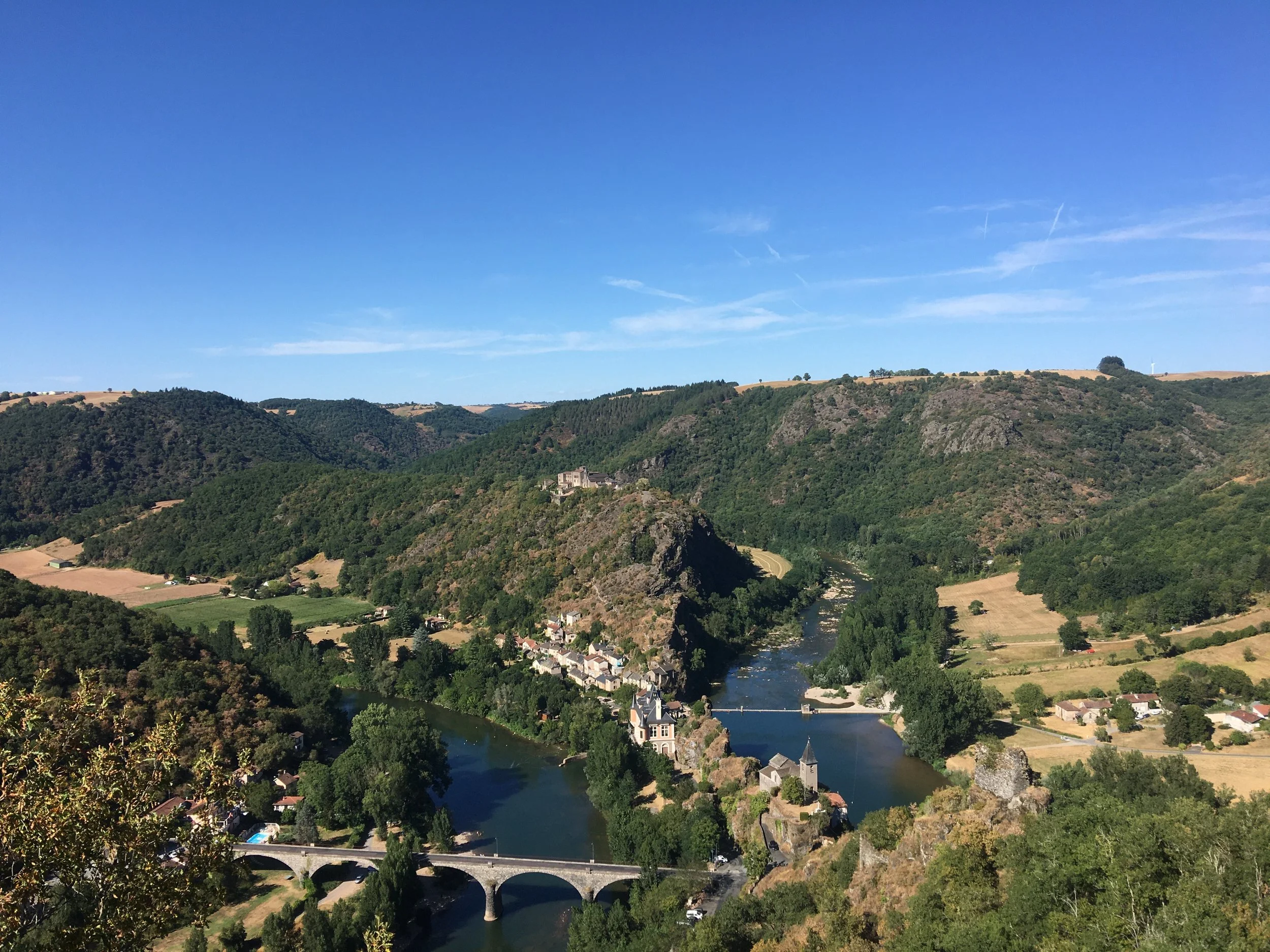 Paysage de vallée avec rivière, ponts, collines verdoyantes et villages sous un ciel bleu clair.