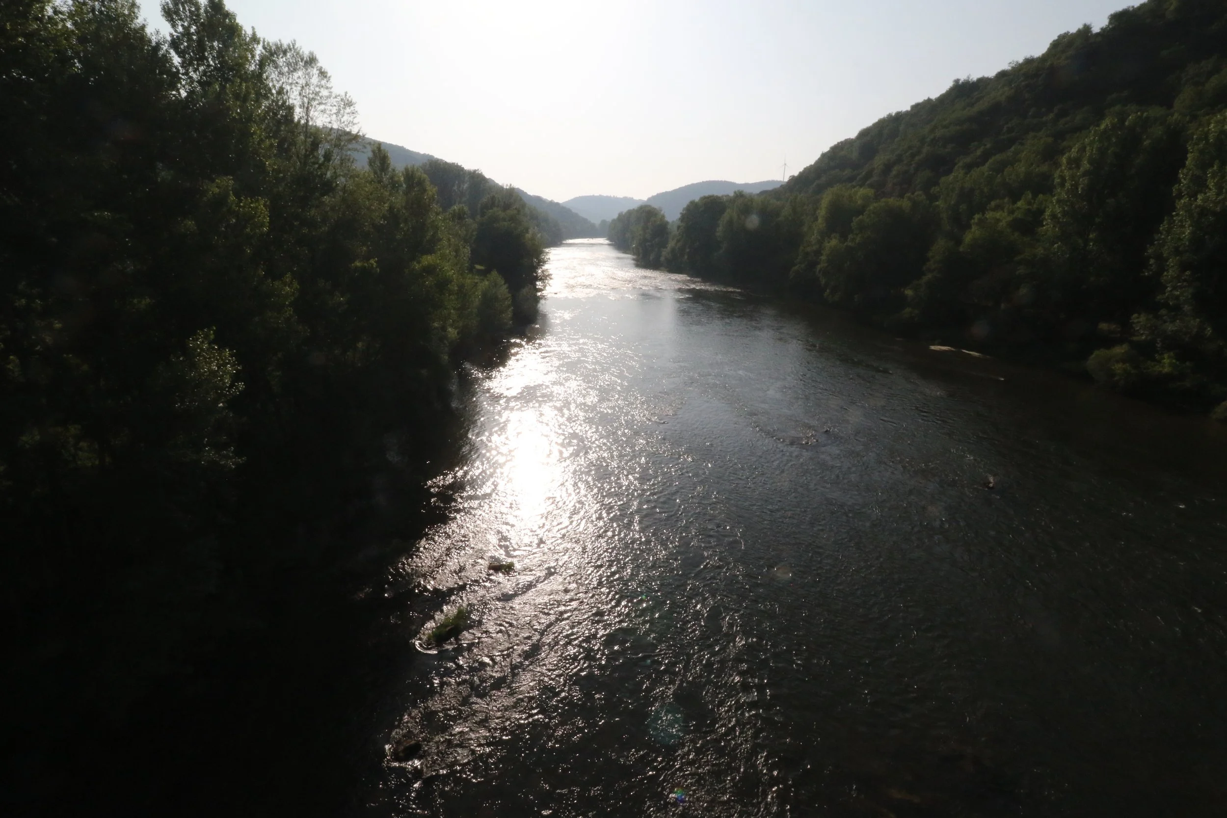 Une rivière bordée d'arbres avec des collines à l'arrière-plan, sous un ciel ensoleillé.