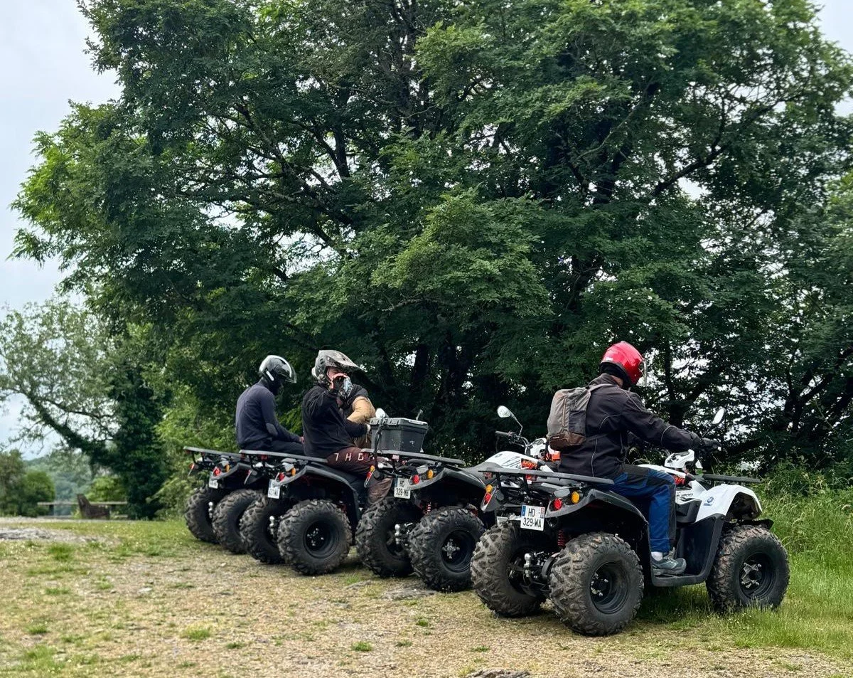 Cinq personnes en équipement de protection, dont deux avec casques complets, assises sur des quads alignés dans un espace vert près d'un arbre.