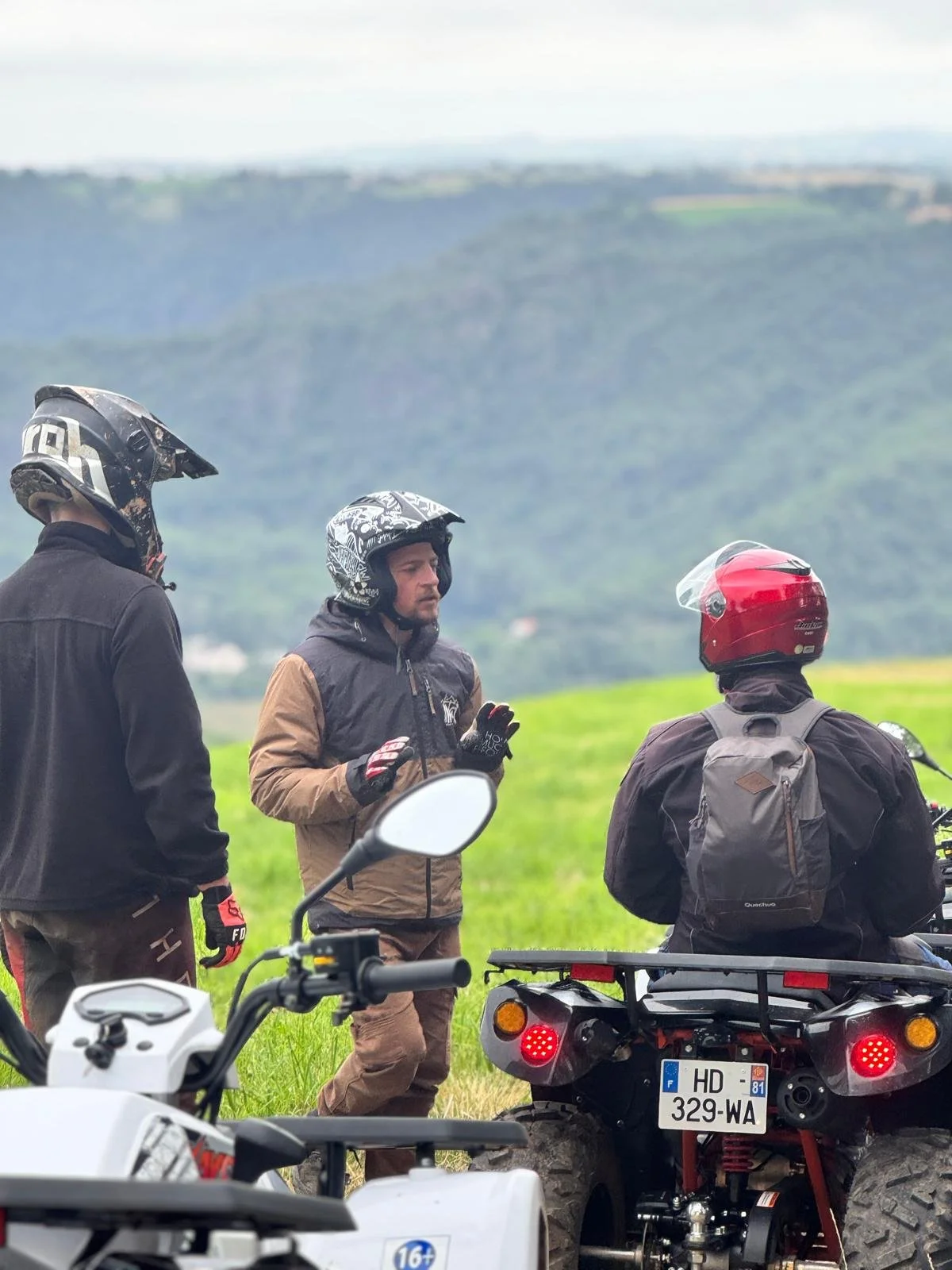 Trois personnes en casques et vêtements de motards, semblent discuter à côté d'une moto quad, dans un champ vert avec une montagne en arrière-plan.