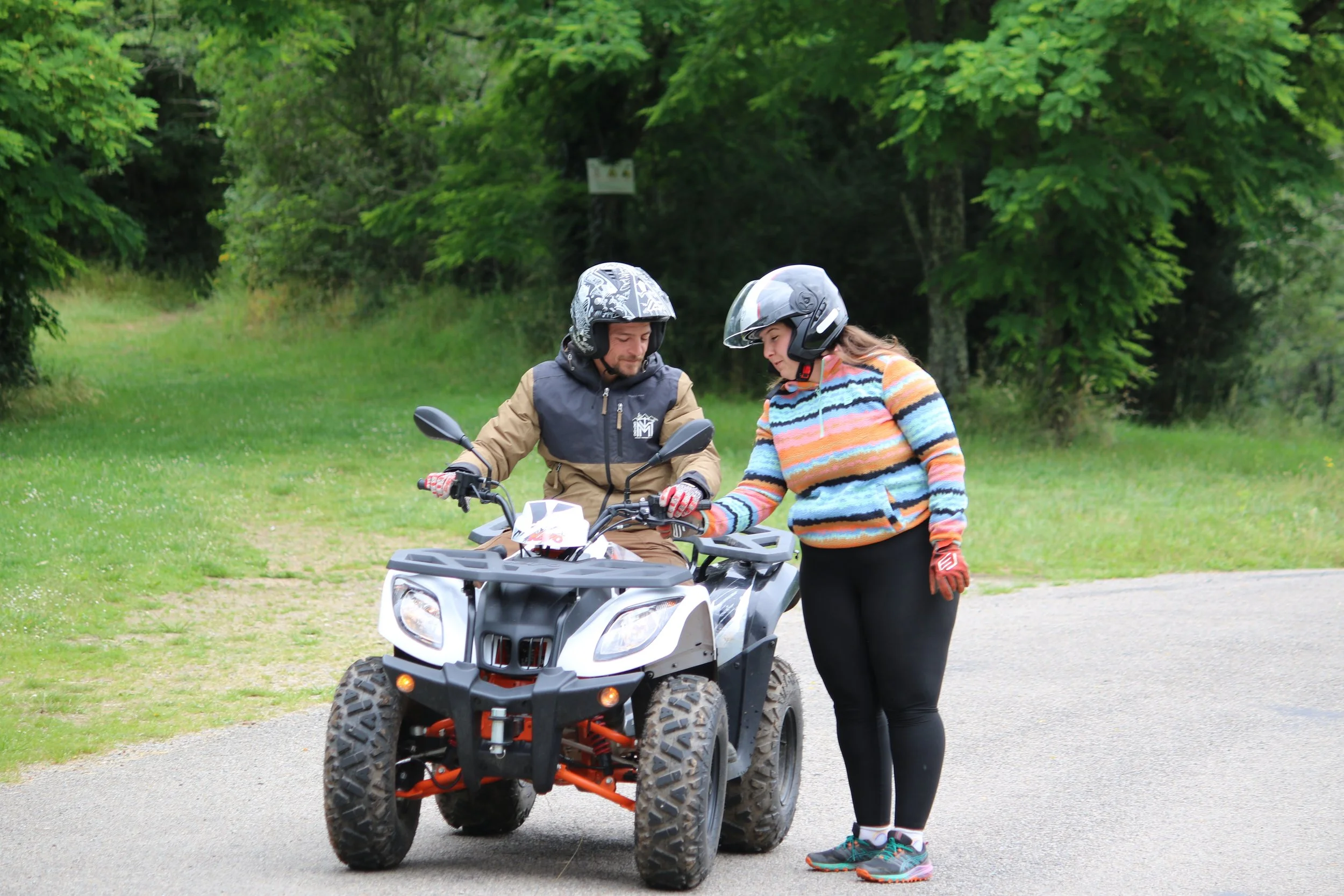 Une femme et un homme portant des casques et des gants discutent à côté d'un quad dans un environnement verdoyant.