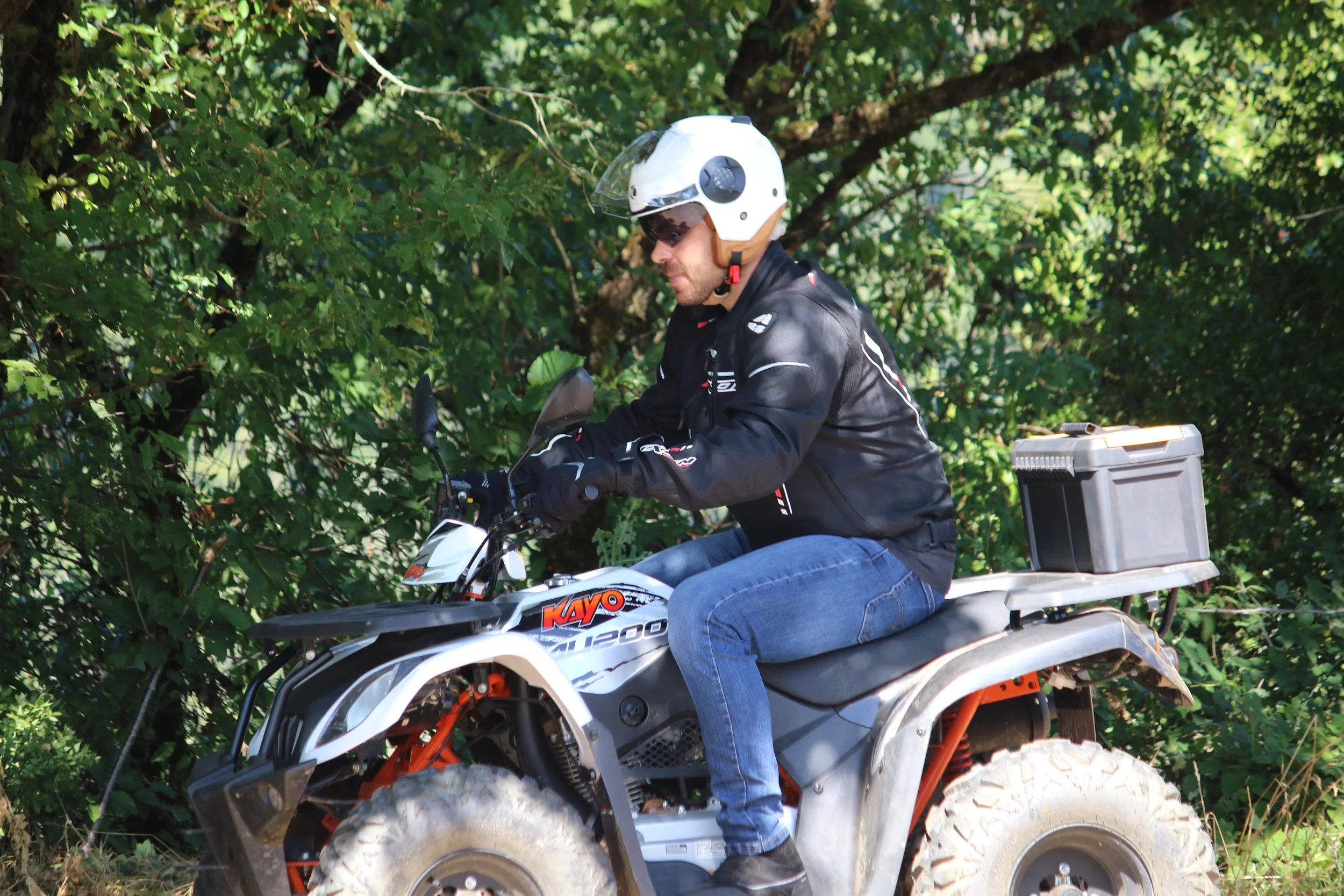 Un homme roulant à moto tout-terrain dans une zone boisée, portant un casque blanc, un blouson noir et des jeans bleus.