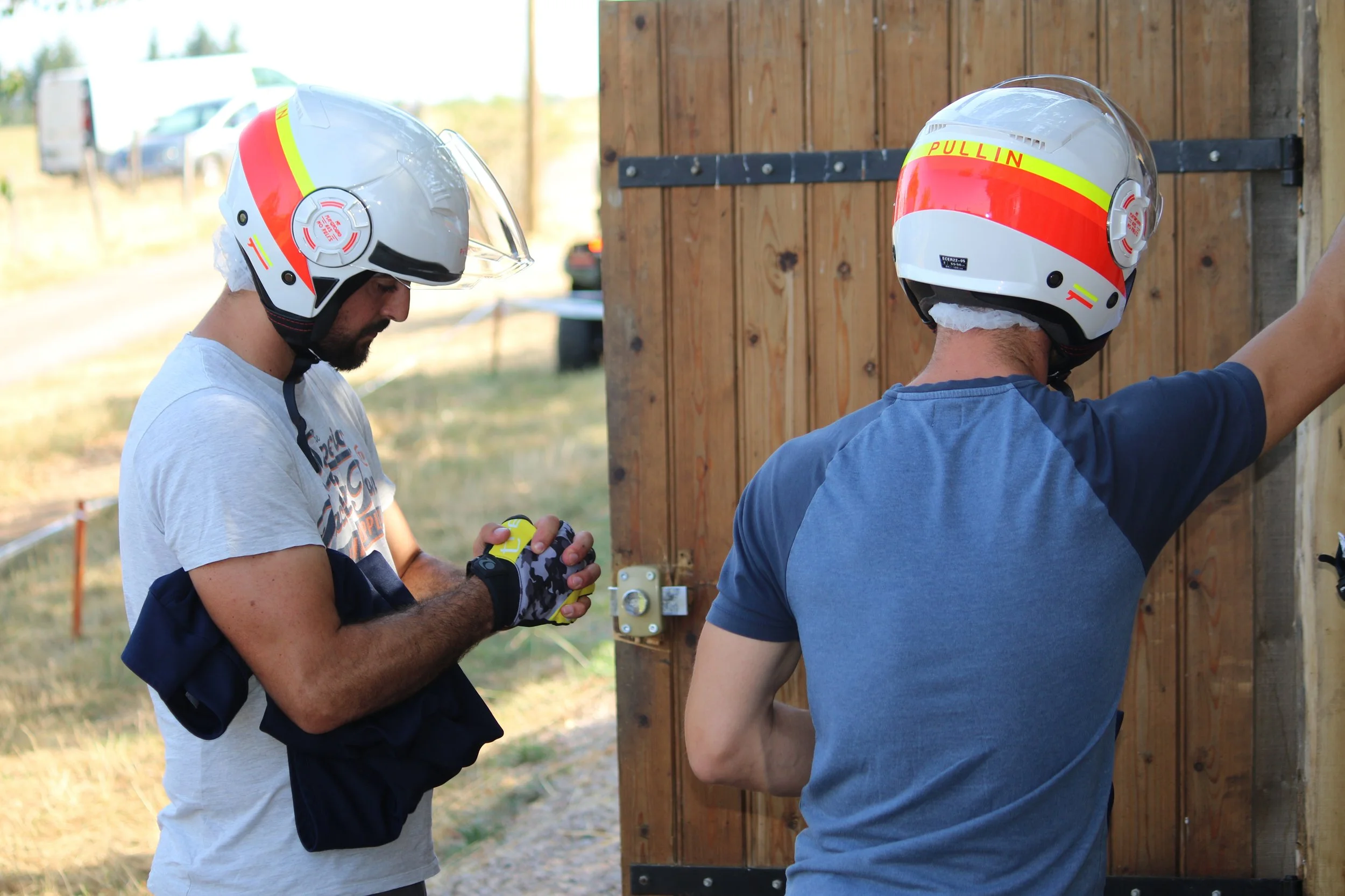 Deux hommes portant des casques de moto près d'une clôture en bois, l'un écrivant quelque chose, l'autre regardant la clôture.