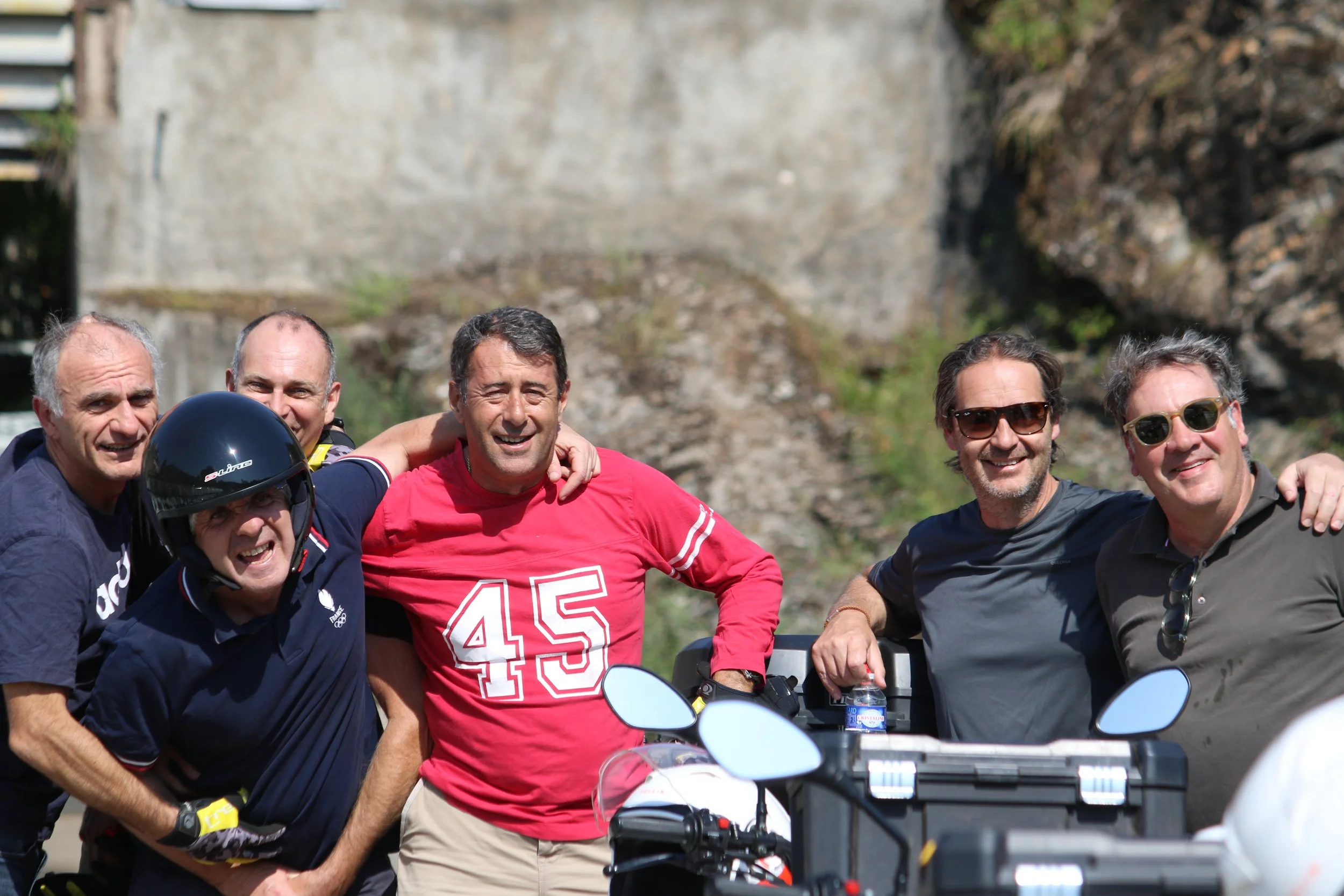 Groupe de six hommes souriants posant ensemble à l'extérieur, certains portant des lunettes de soleil, un portant un casque, avec un arrière-plan rocheux.