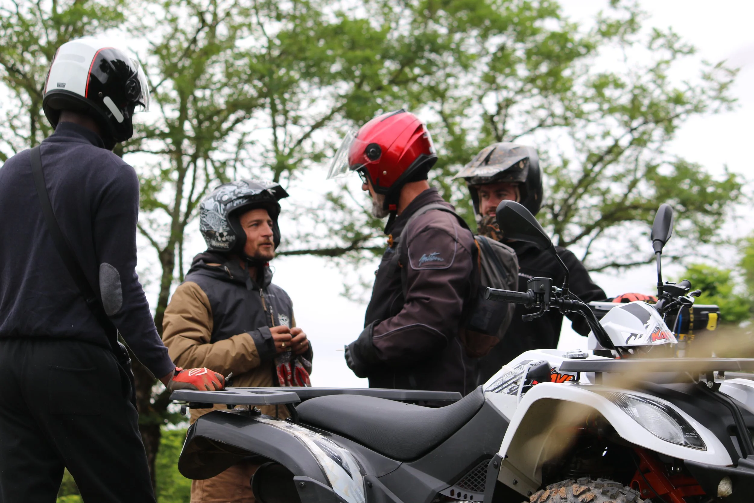 Groupe de quatre personnes avec casques de moto, discutant à côté d'une moto tout-terrain en extérieur avec des arbres en arrière-plan.