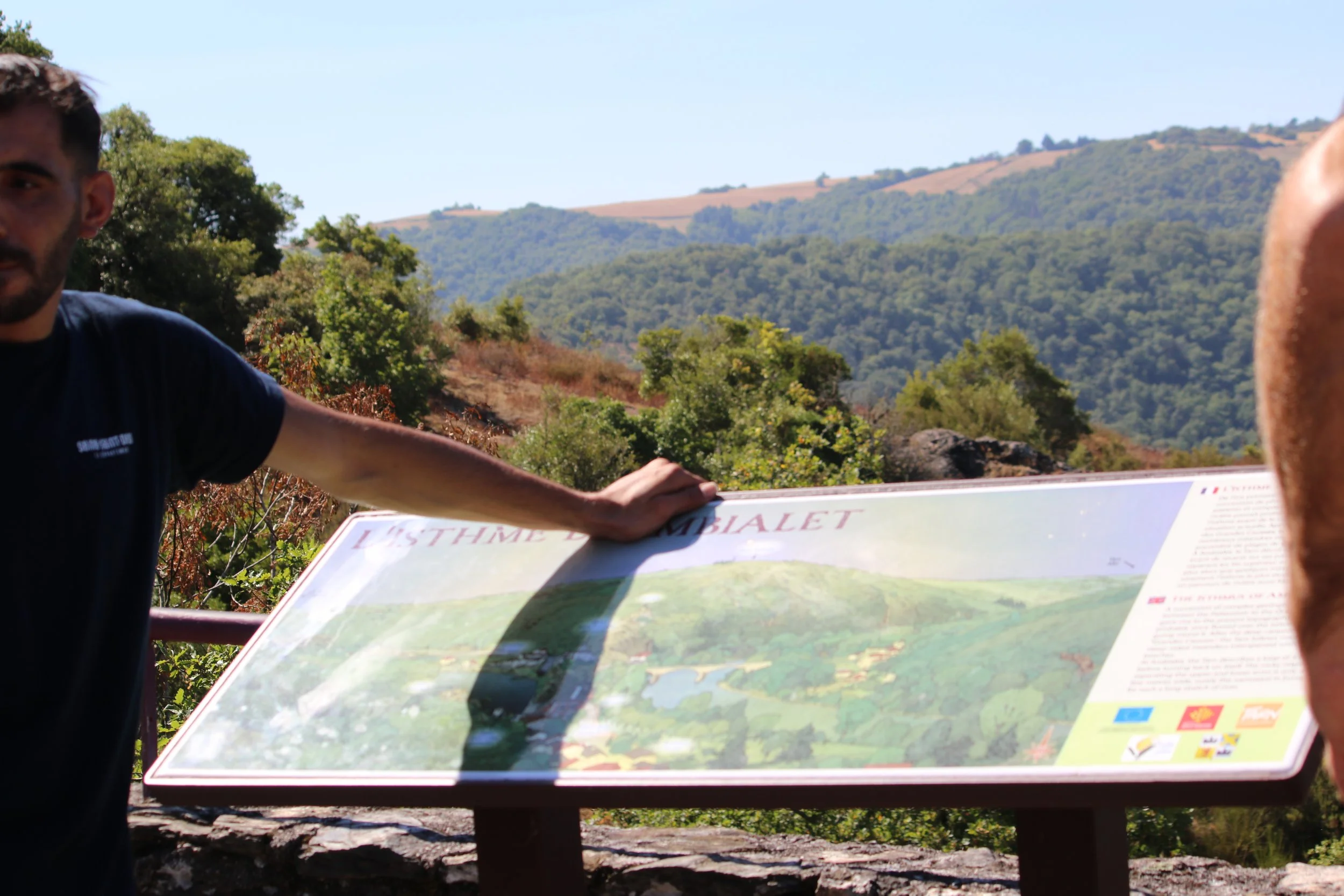 Un homme se tenant devant une plaque d'information touristique avec un paysage de collines verdoyantes en arrière-plan, en plein air par temps ensoleillé.