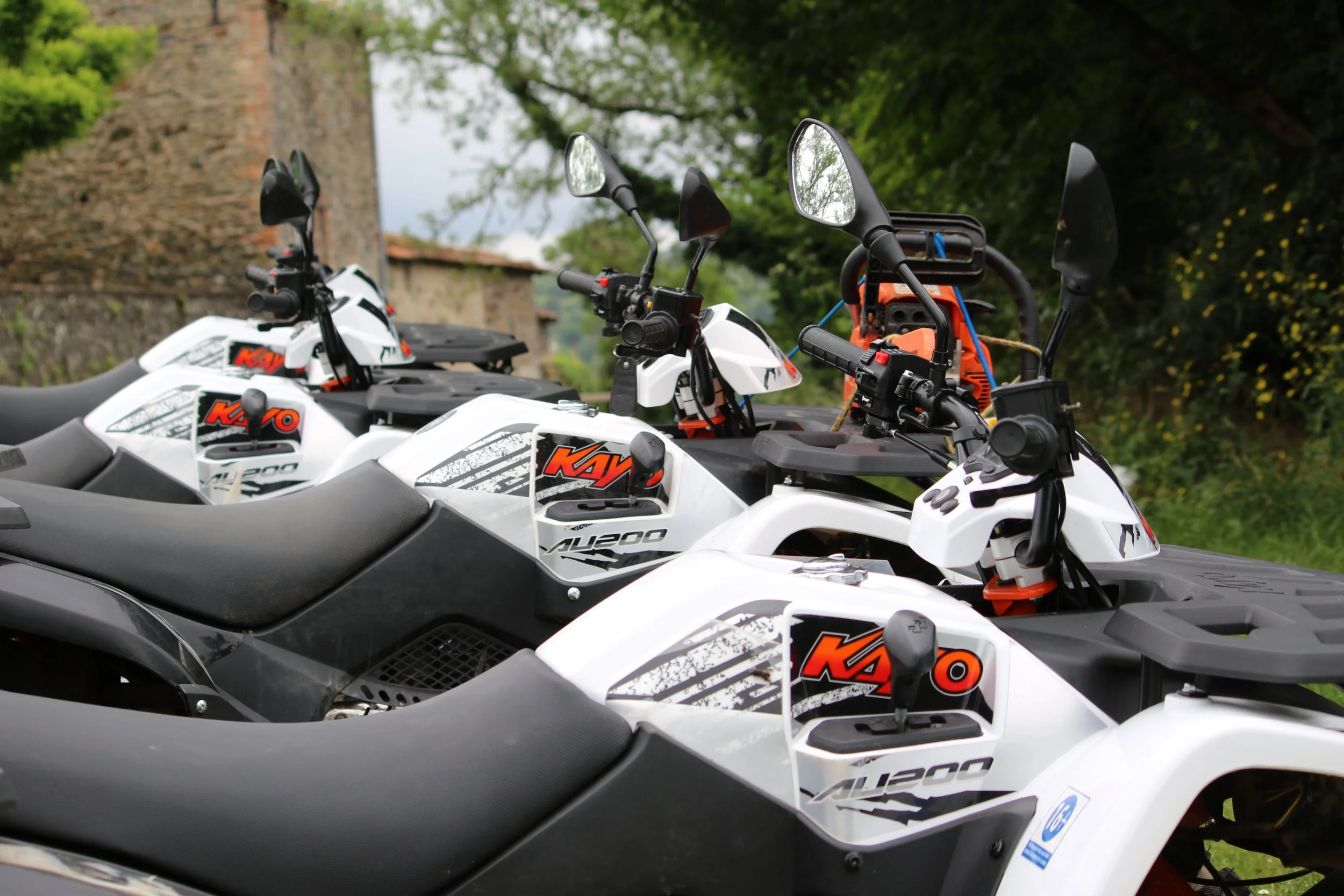 Trois quads tout terrain de couleur blanche et noire, alignés en rangée dans un environnement en plein air avec des arbres verts et un vieux mur en pierre en arrière-plan.