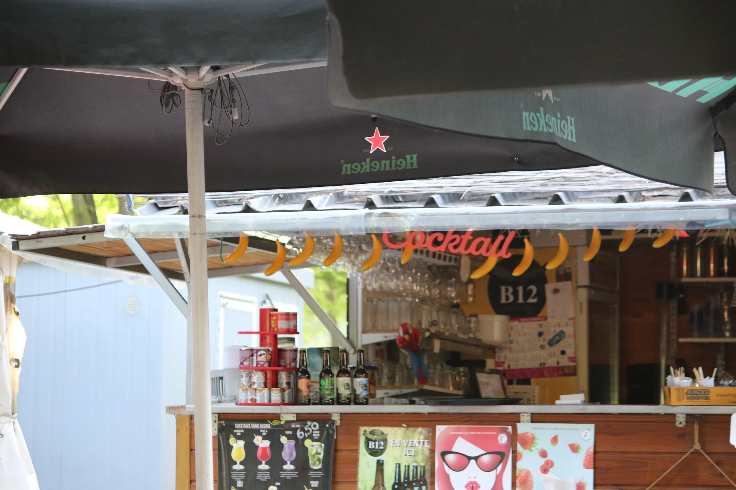 Bar à cocktails avec un panneau indiquant 'Cocktail', décoré de bananes suspendues, présente des bouteilles, des canettes, des verres suspendus, et un menu avec différentes options de cocktails.