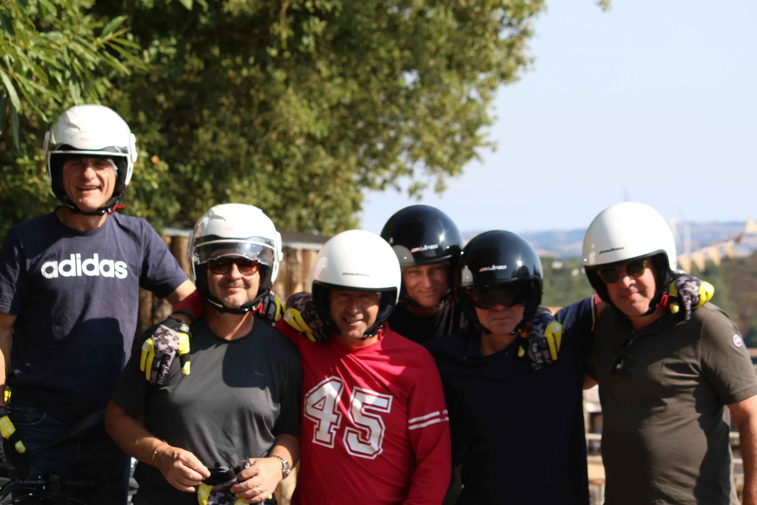 Groupe de six hommes portant des casques de moto, certains avec lunettes de soleil, souriants, avec des arbres en arrière-plan.