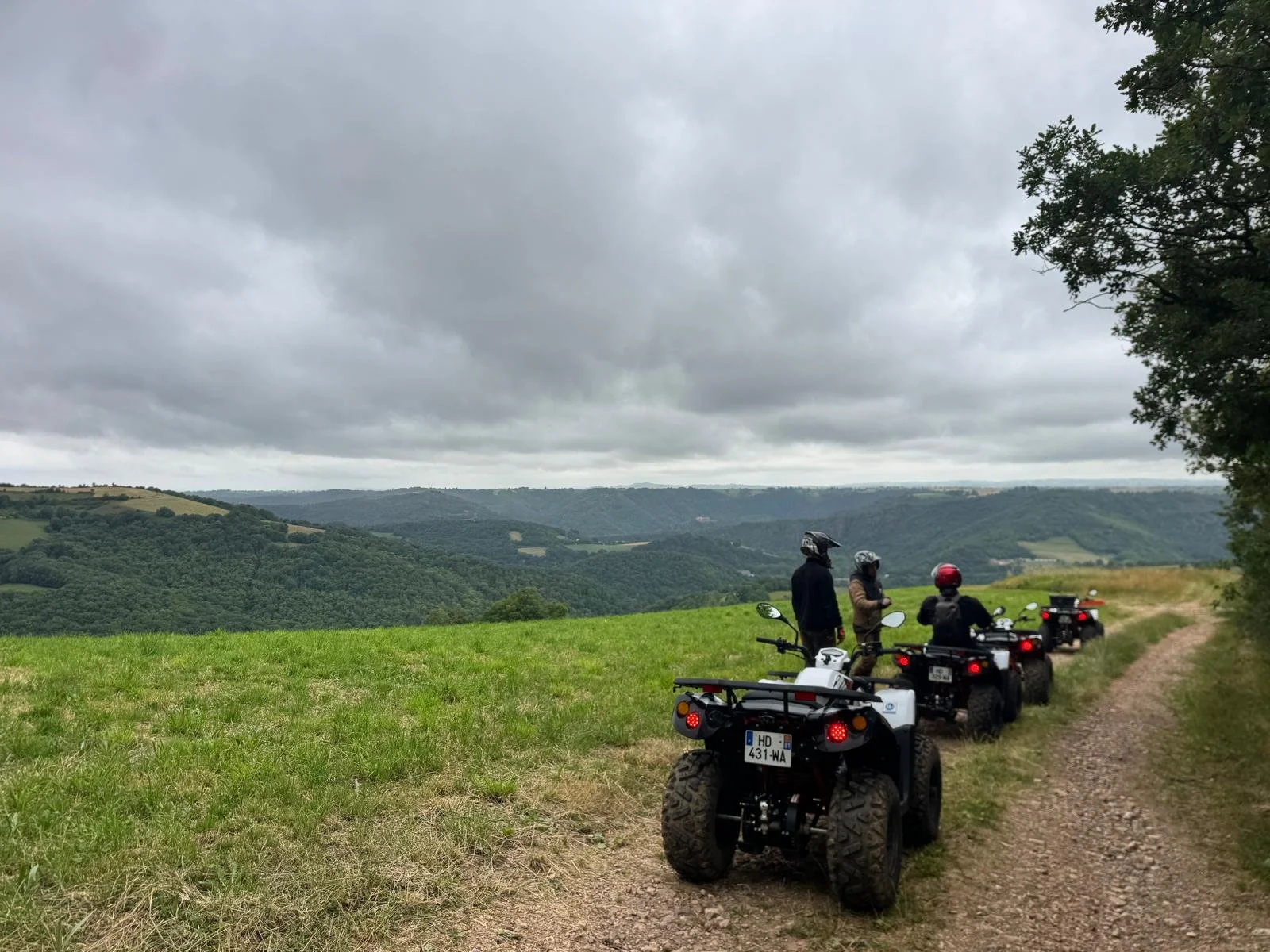 Groupe de quatre personnes en casques, assises sur des quads, regardant un paysage de collines verdoyantes sous un ciel nuageux.