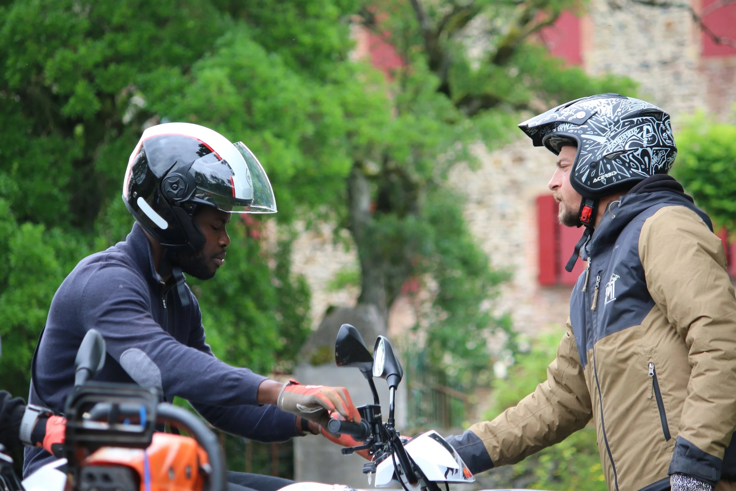 Deux hommes en casque de moto échangent un document ou une carte, entourés de verdure.