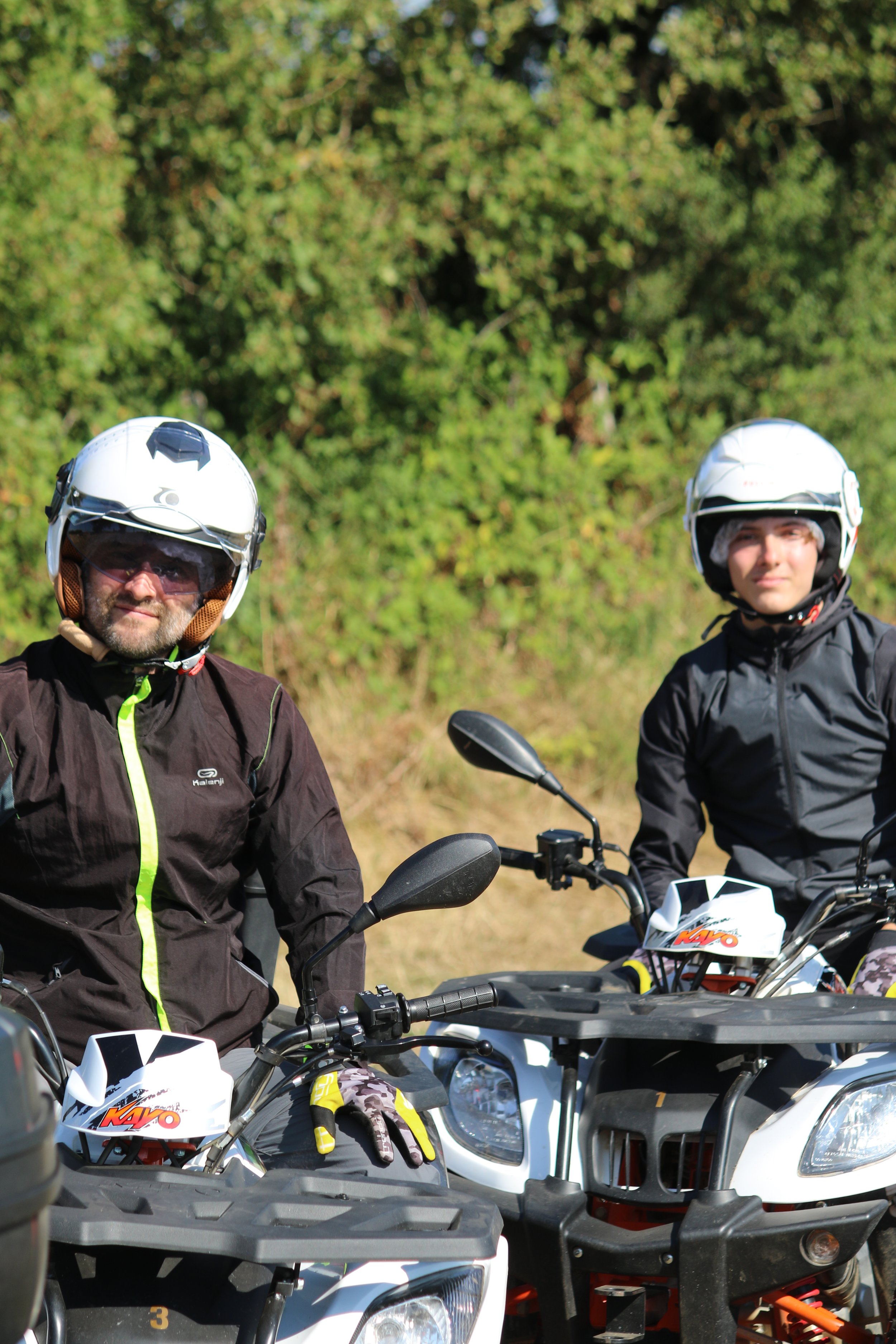 Deux personnes en casques et vêtements de moto tout-terrain, assises sur des quad, en plein air avec des arbres en arrière-plan.
