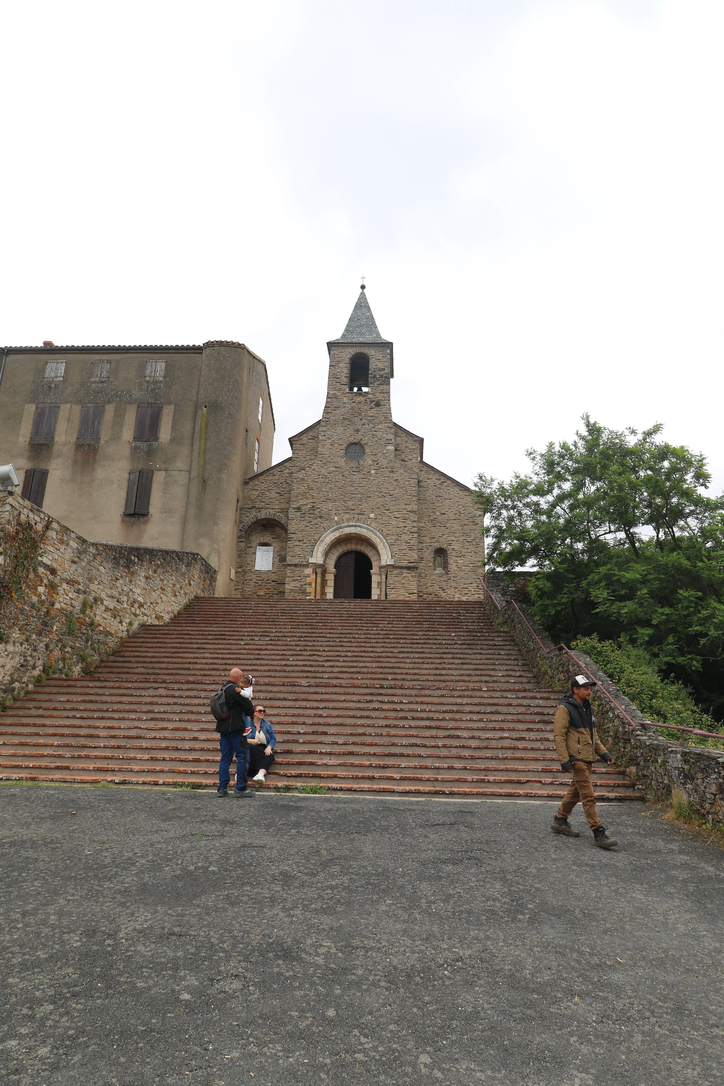 Une église en pierre avec un escalier en pierre menant à l'entrée, entourée d'arbres, avec trois personnes devant l'église, deux debout et une assise, sous un ciel nuageux.