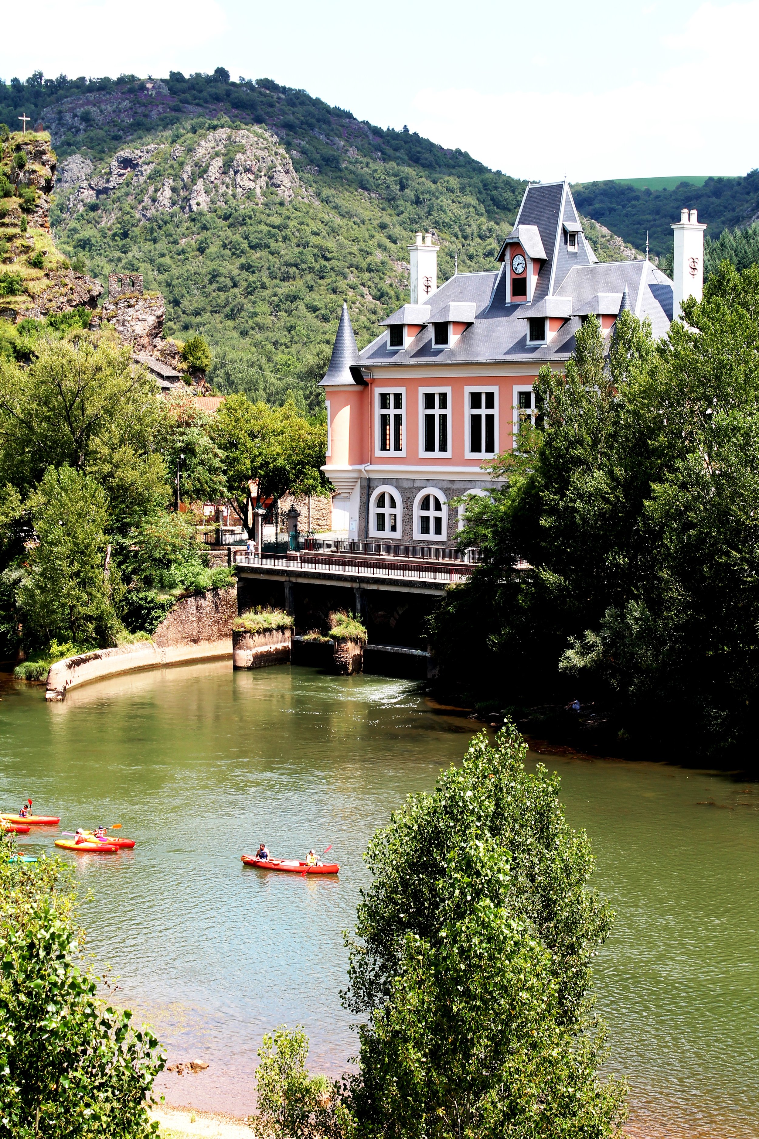 Une maison de style victorien rose avec un toit gris, située au bord d'une rivière entourée de verdure, avec des montagnes en arrière-plan.