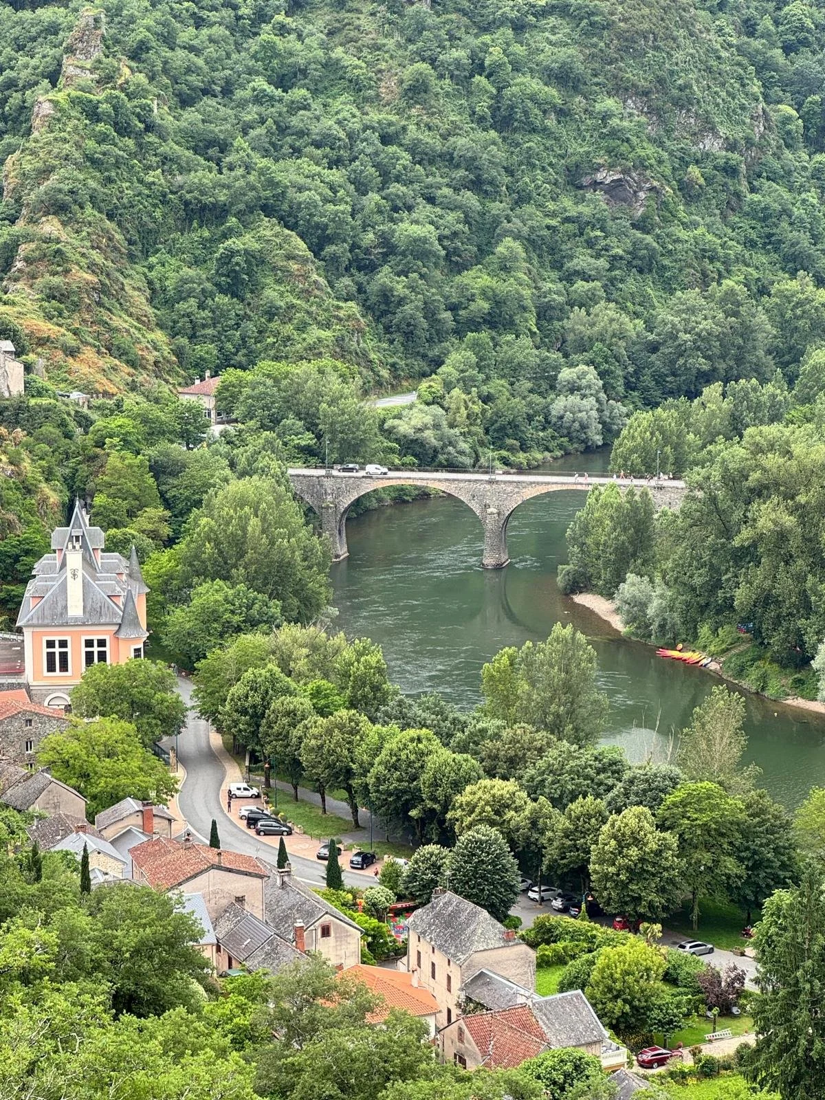 Village avec des maisons en toit en tuiles rouges au bord d'une rivière, entouré de nombreux arbres verts, avec un pont en pierre traversant la rivière et une forêt montagneuse en arrière-plan.