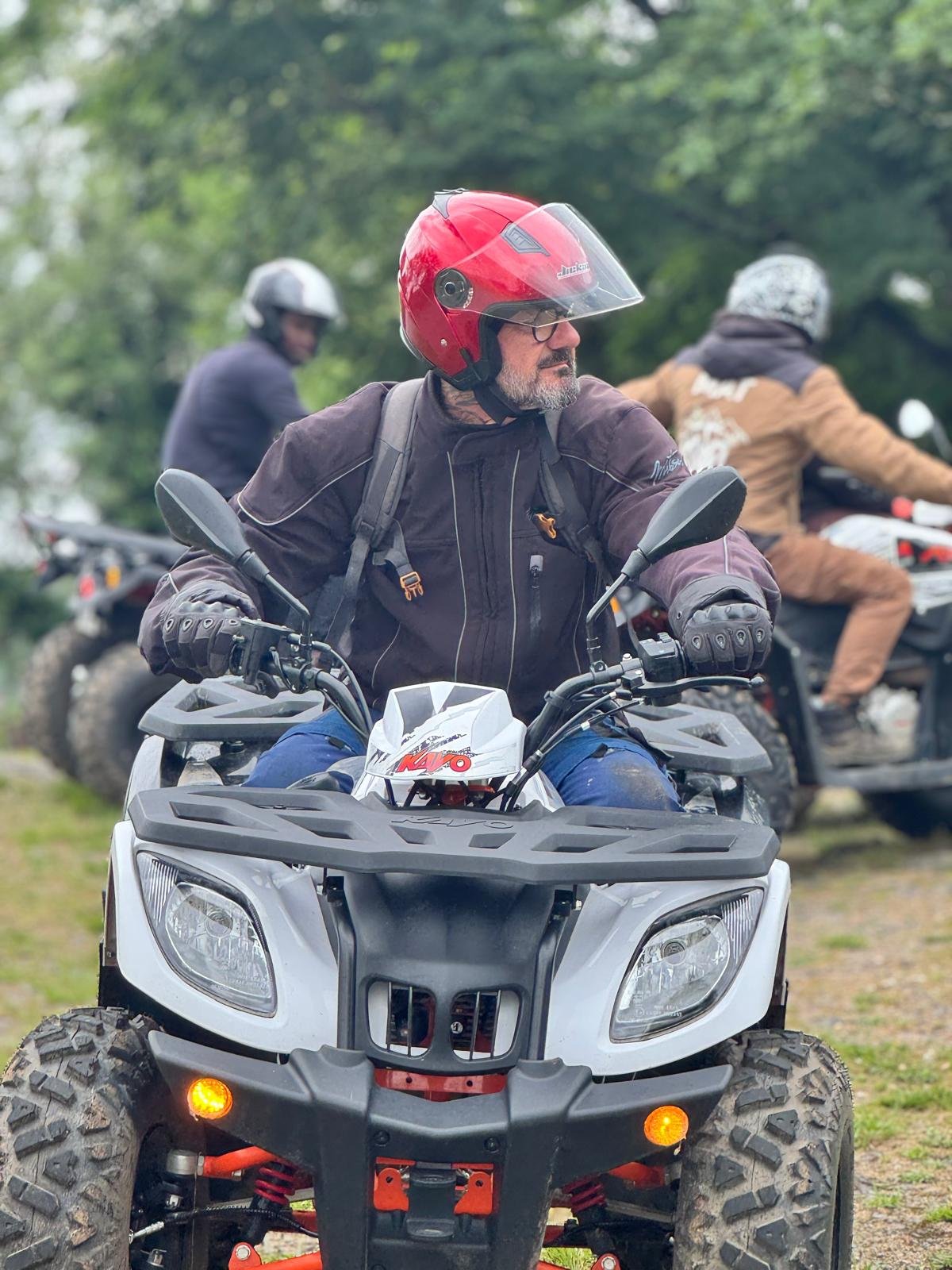 Un homme portant un casque rouge et une veste noire est assis sur un quad, avec deux autres personnes en arrière-plan également sur des quads. La scène se déroule en plein air avec des arbres en arrière-plan.