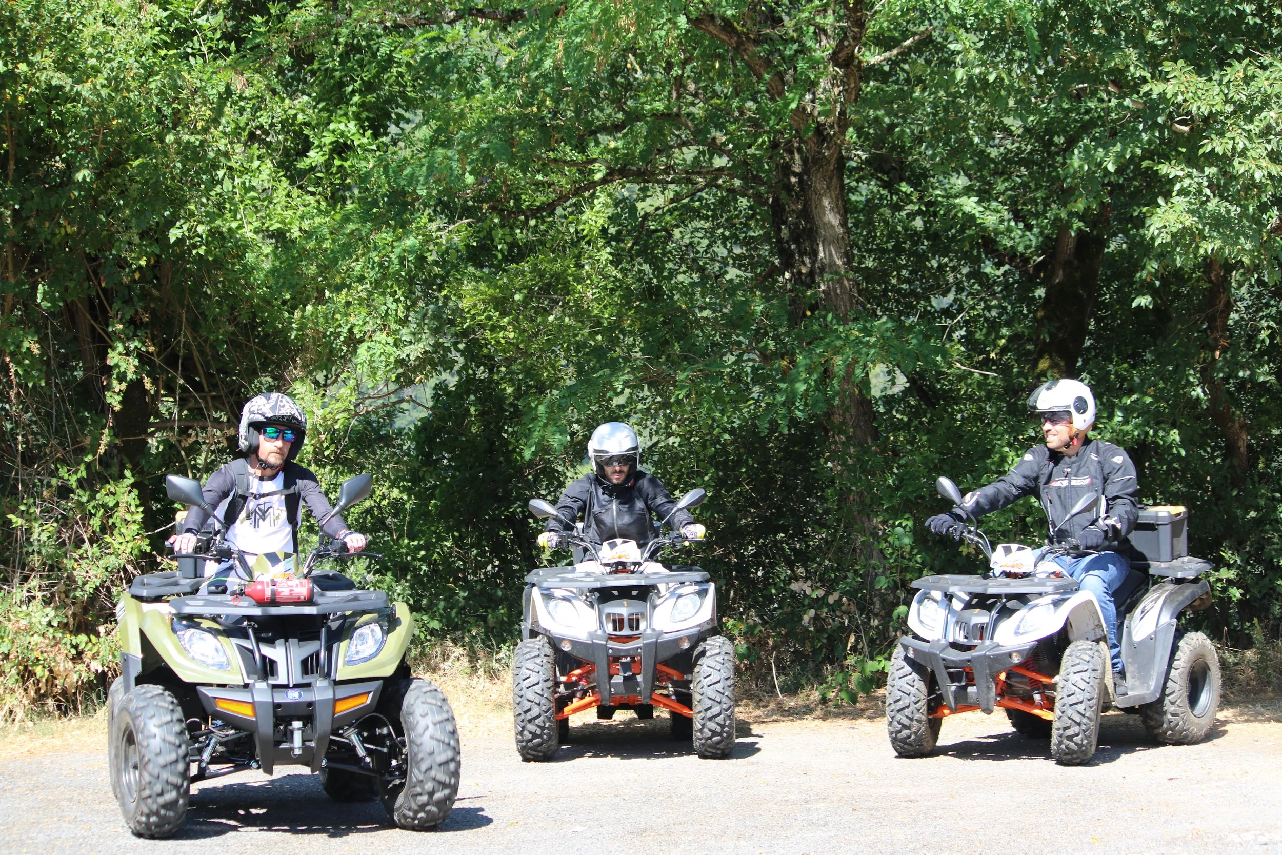 Trois personnes en casques et tenues de moto tout-terrain en position de conduite, assises sur des quads tout-terrain, devant un arbre en forêt.