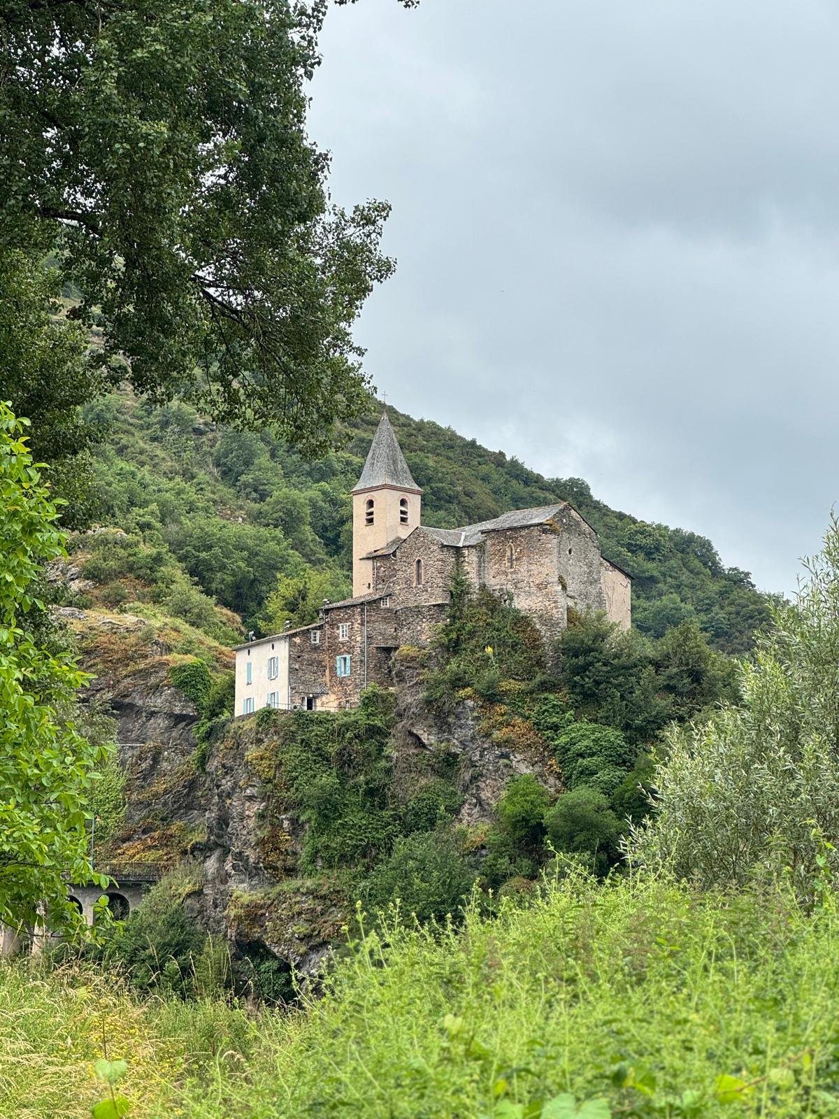 Une vieille église en pierre perchée sur une falaise entourée de verdure et de montagnes, sous un ciel nuageux.