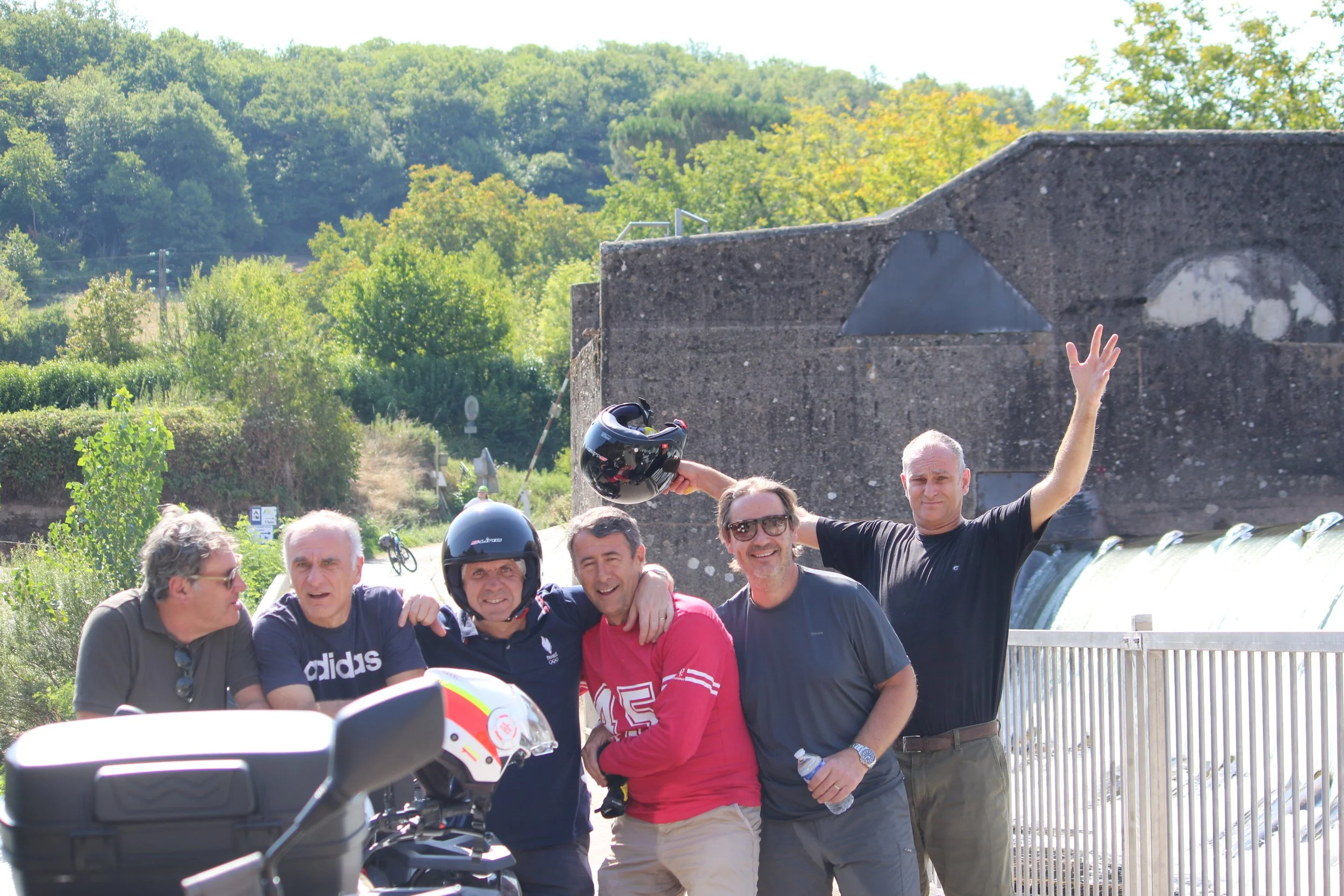 groupe de six hommes souriants posant ensemble, certains portant des casques de moto, en plein air avec une nature verdoyante en arrière-plan.