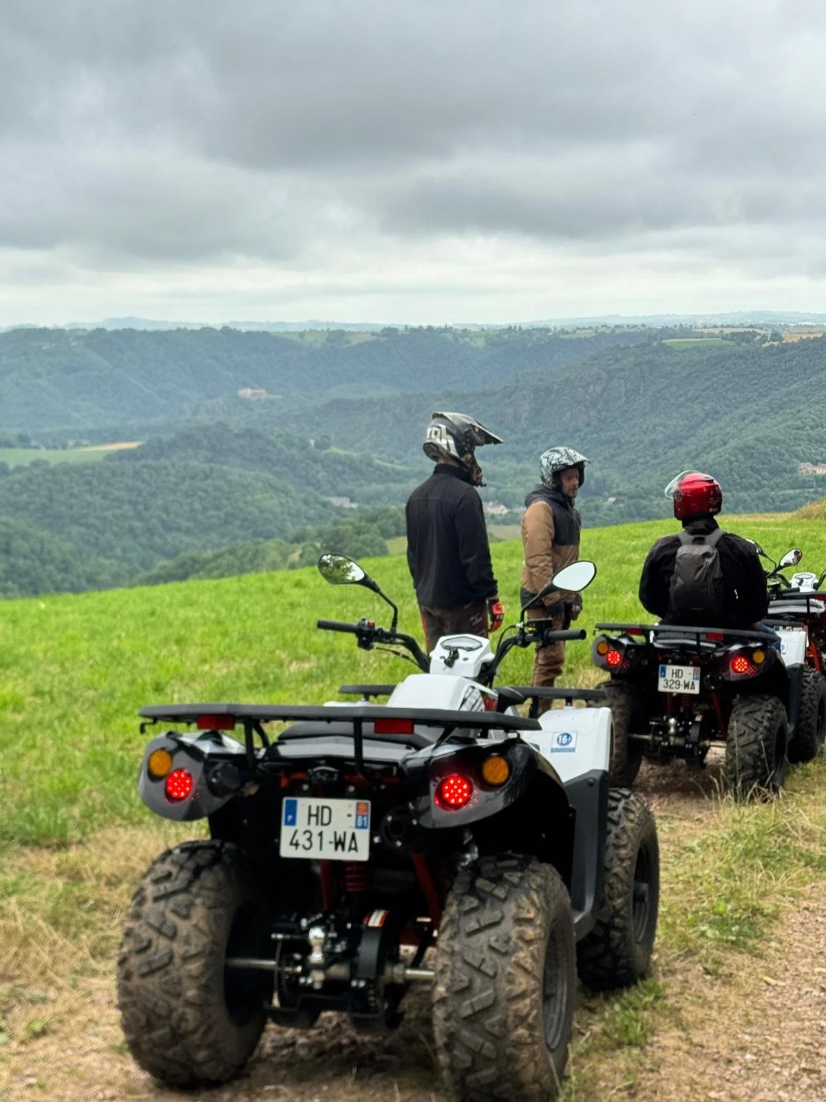 Trois motocyclistes avec casques se reposent dans un champ avec une vue panoramique de montagnes et de vallées sous un ciel nuageux.