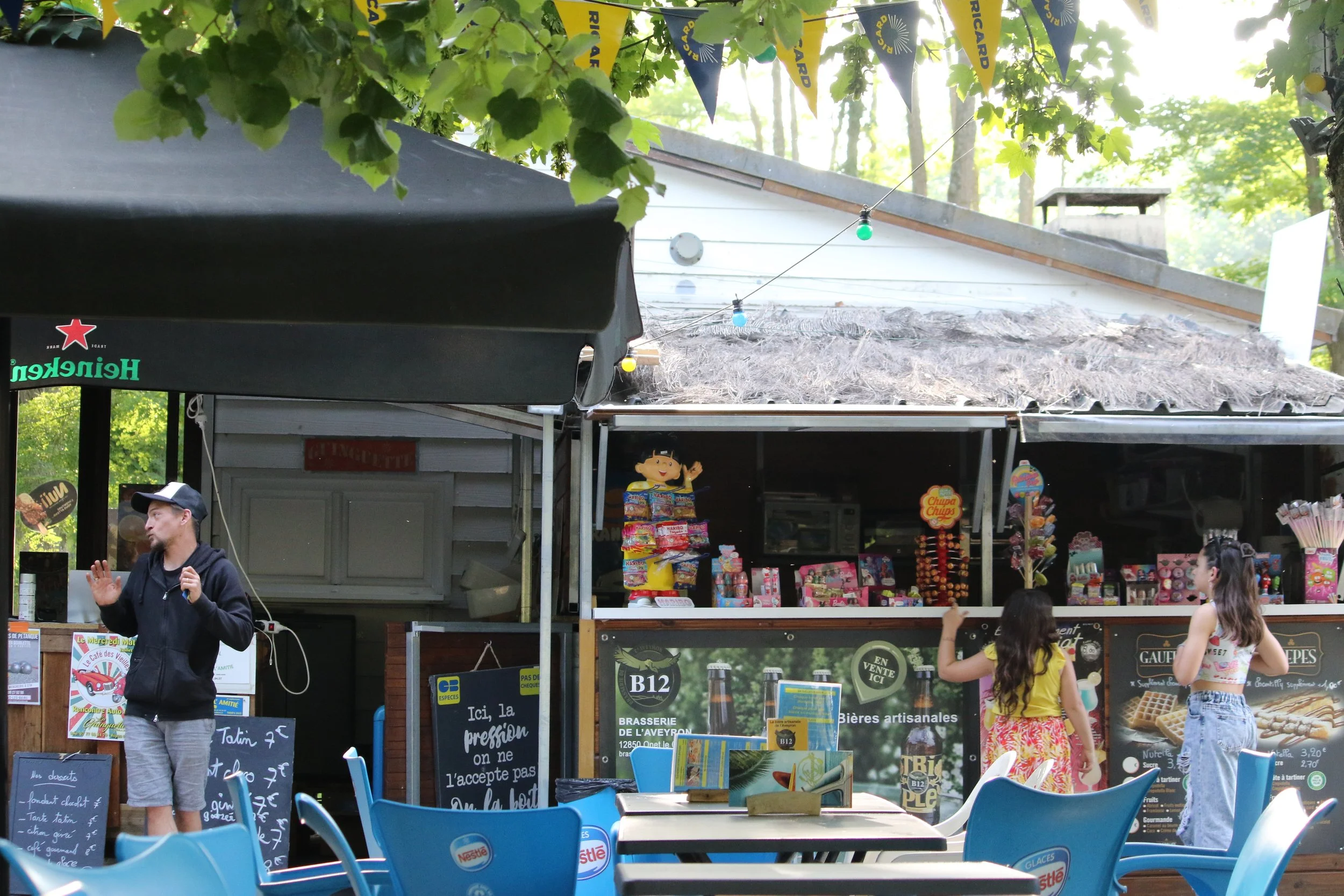 Stand de vente de confiseries avec deux jeunes filles à proximité, un homme parlant avec un micro à côté, tables vides en premier plan, parasol Heineken, et décorations de fête foraine en arrière-plan.