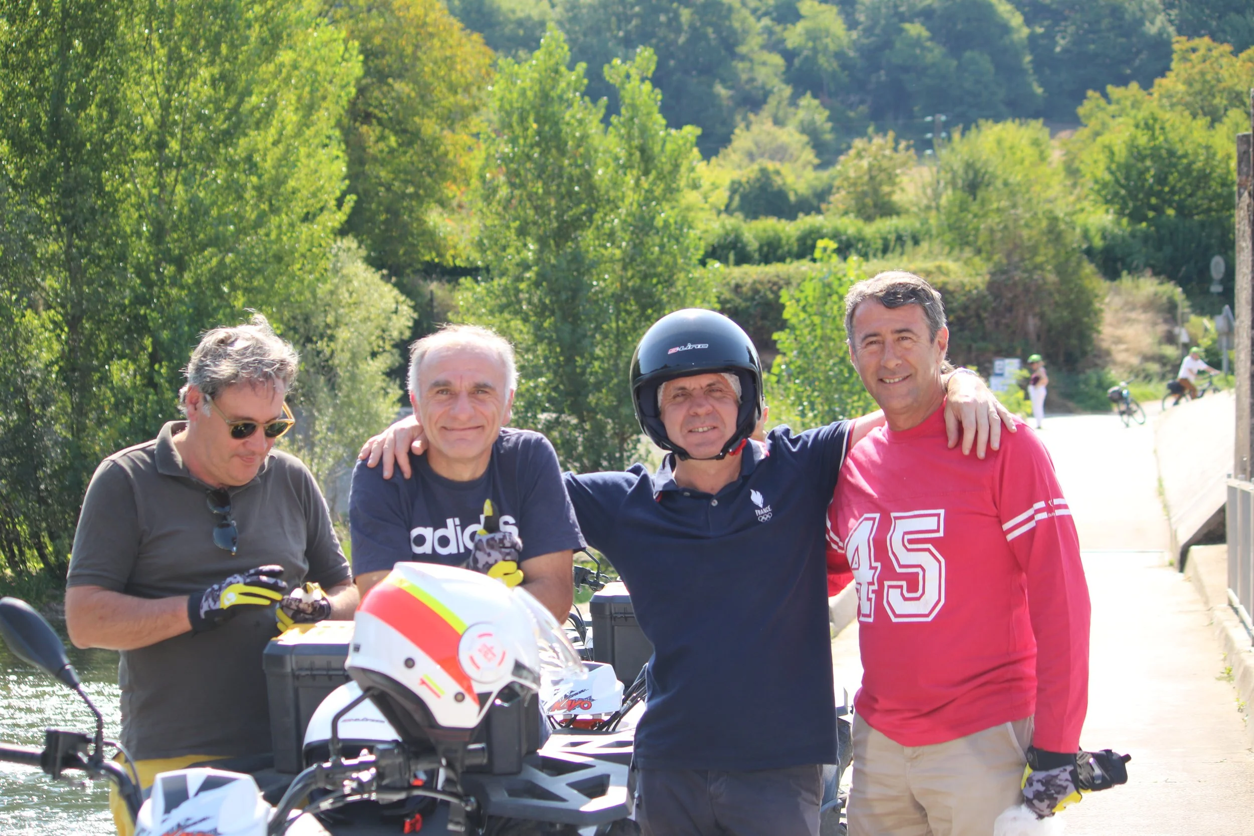 Quatre hommes en plein air près d'une rivière, deux d'entre eux portant des casques de moto, souriant et posant pour la photo, avec des arbres verts en arrière-plan.