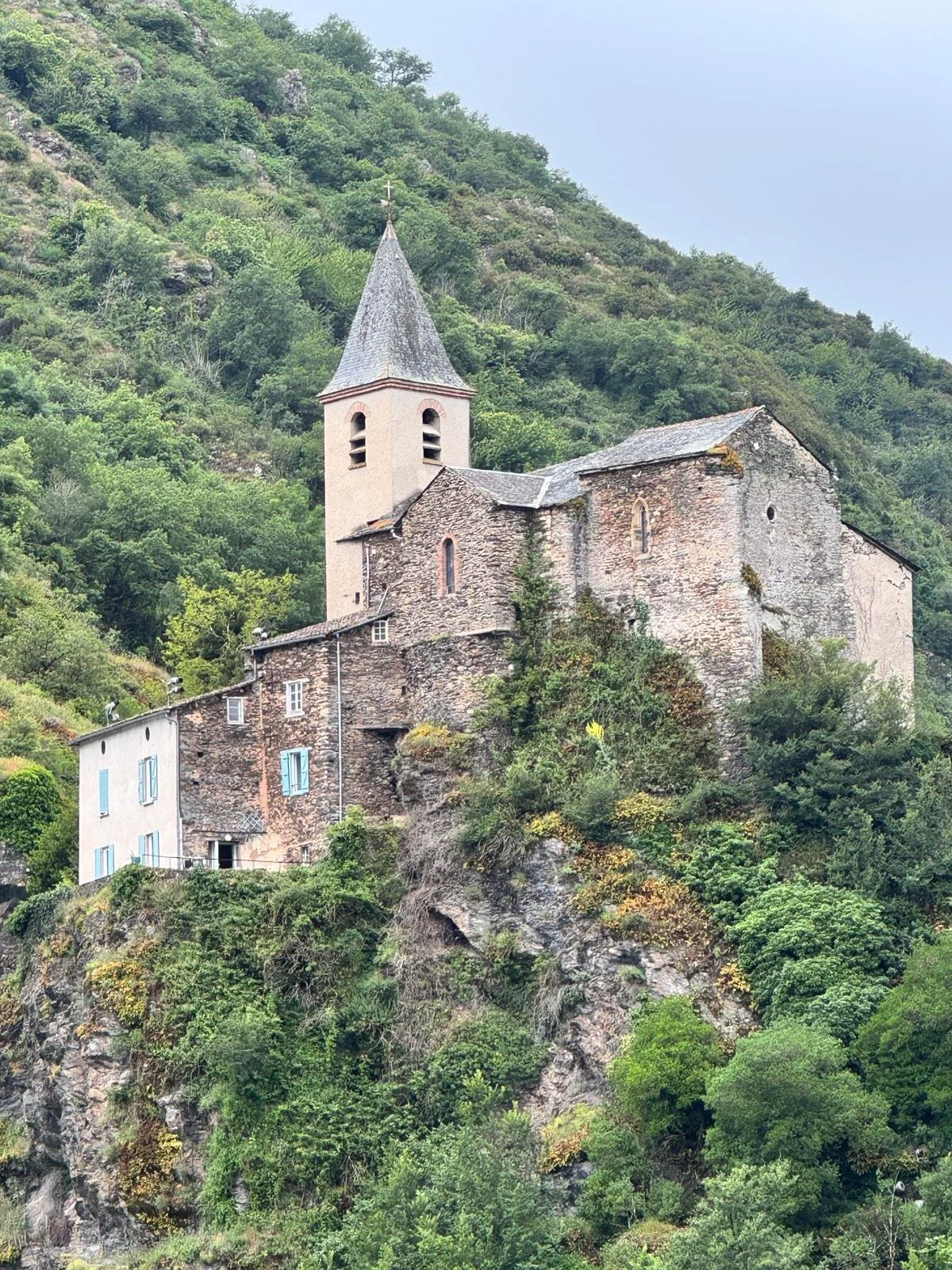 Une vieille église en pierre, perchée sur une falaise escarpée couverte de verdure, avec un clocher pointu et une croix au sommet.