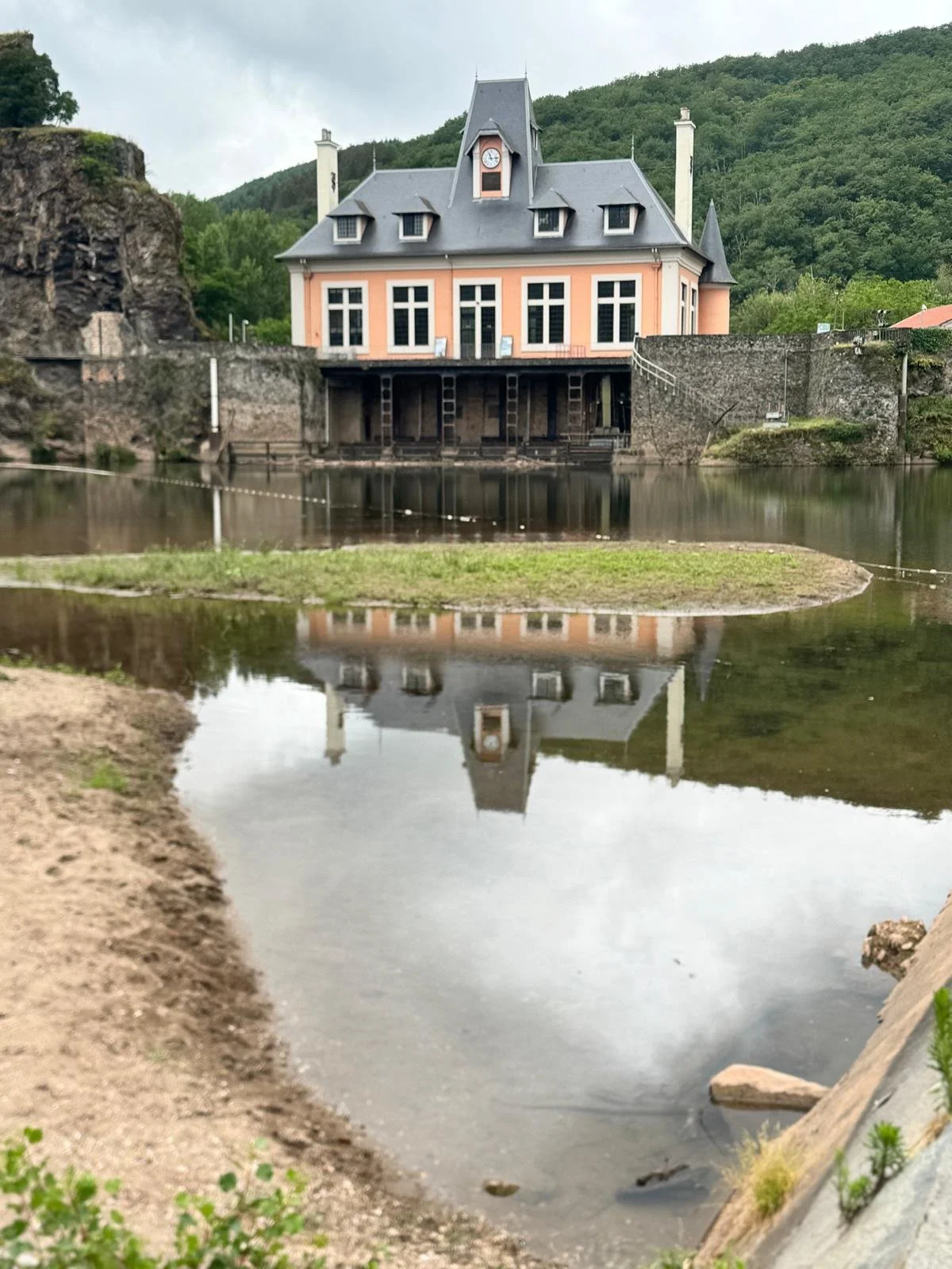 Une maison de style victorien avec un toit en pente, située au bord d'un lac, avec des montagnes vertes en arrière-plan, et une reflection dans l'eau du lac.