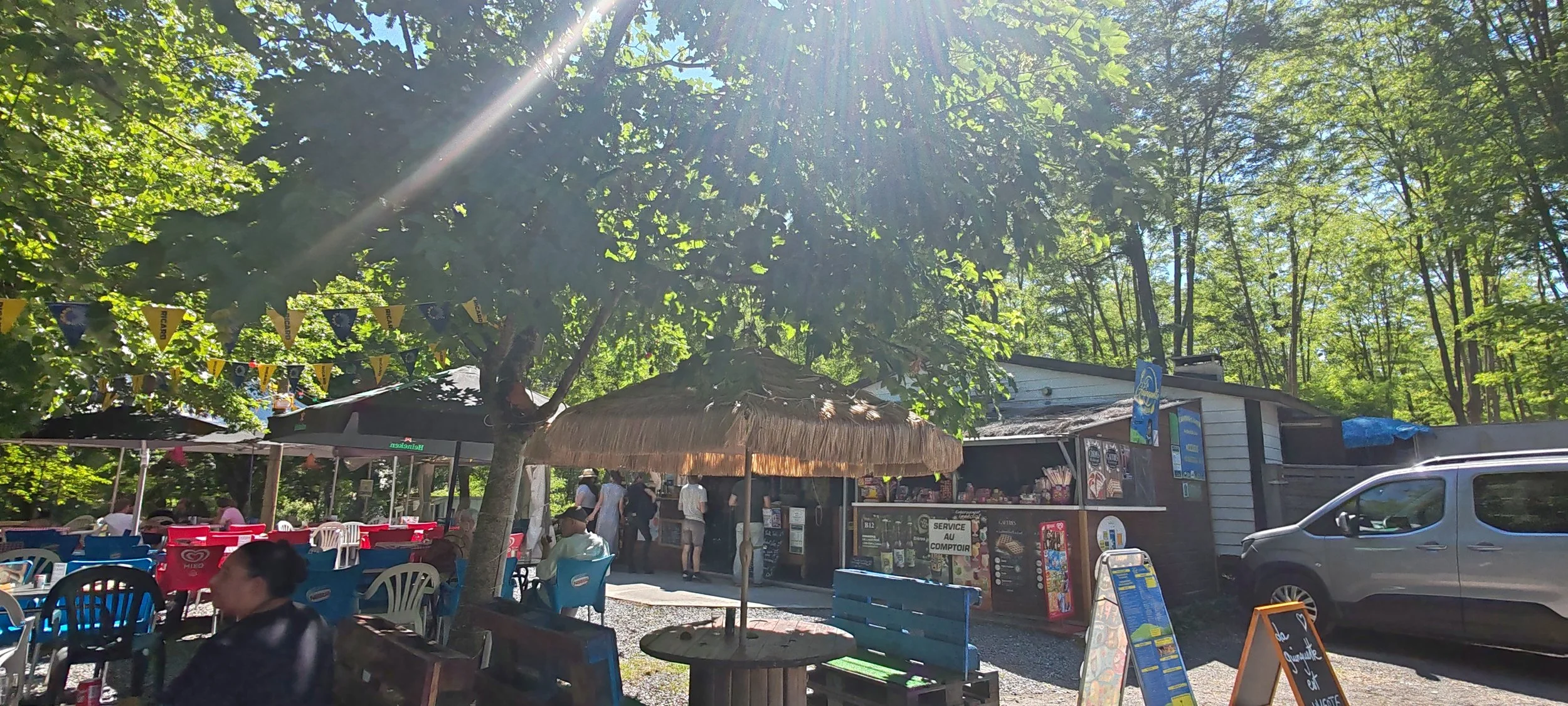Terrasse en plein air d'un café ou restaurant dans une forêt au soleil, avec tables, chaises colorées, parasols, un kiosque ou point de vente, et des personnes profitant de l'ambiance.
