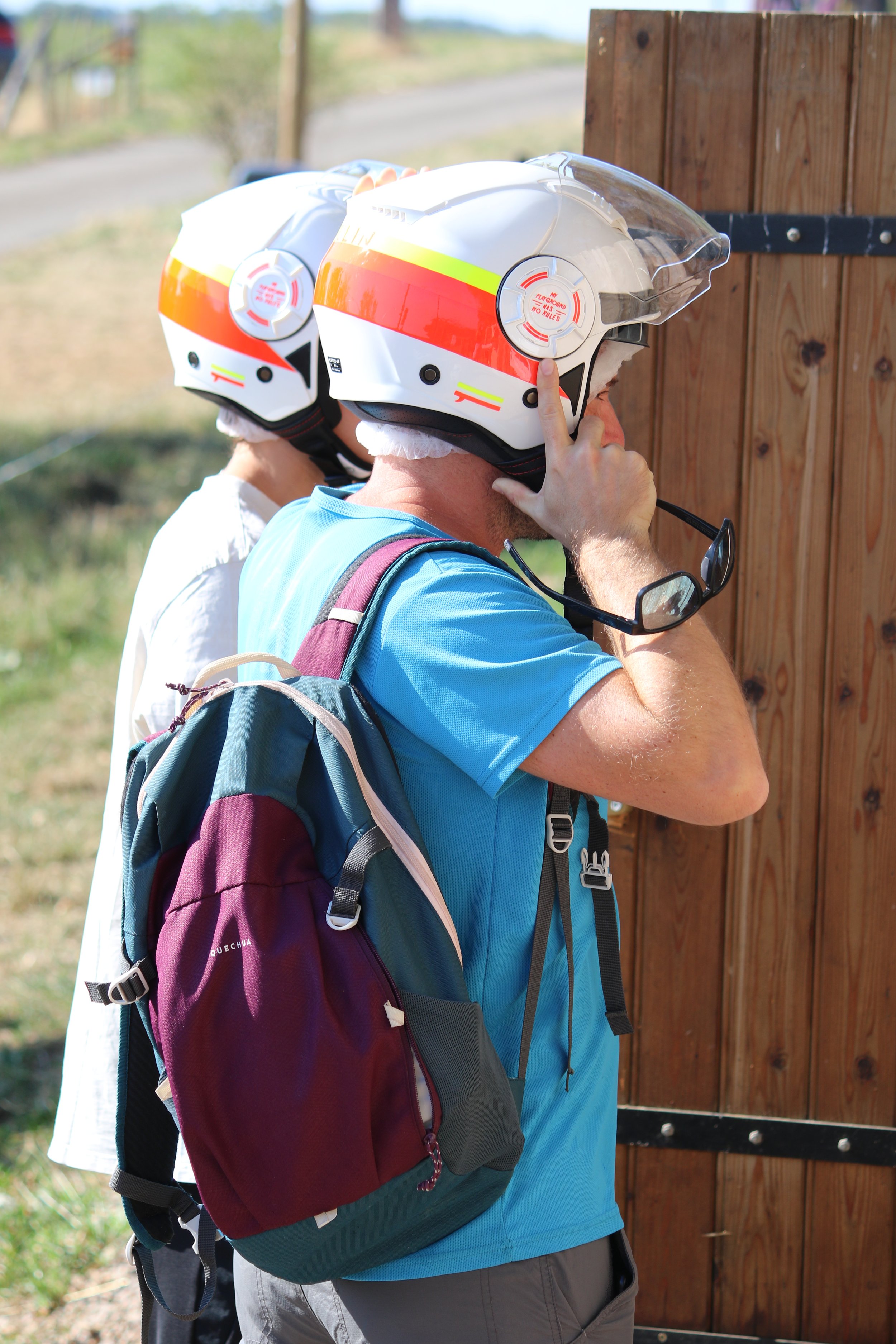 Deux personnes portant un casque de moto, l'une ajustant son casque, à côté d'une clôture en bois, en plein air.