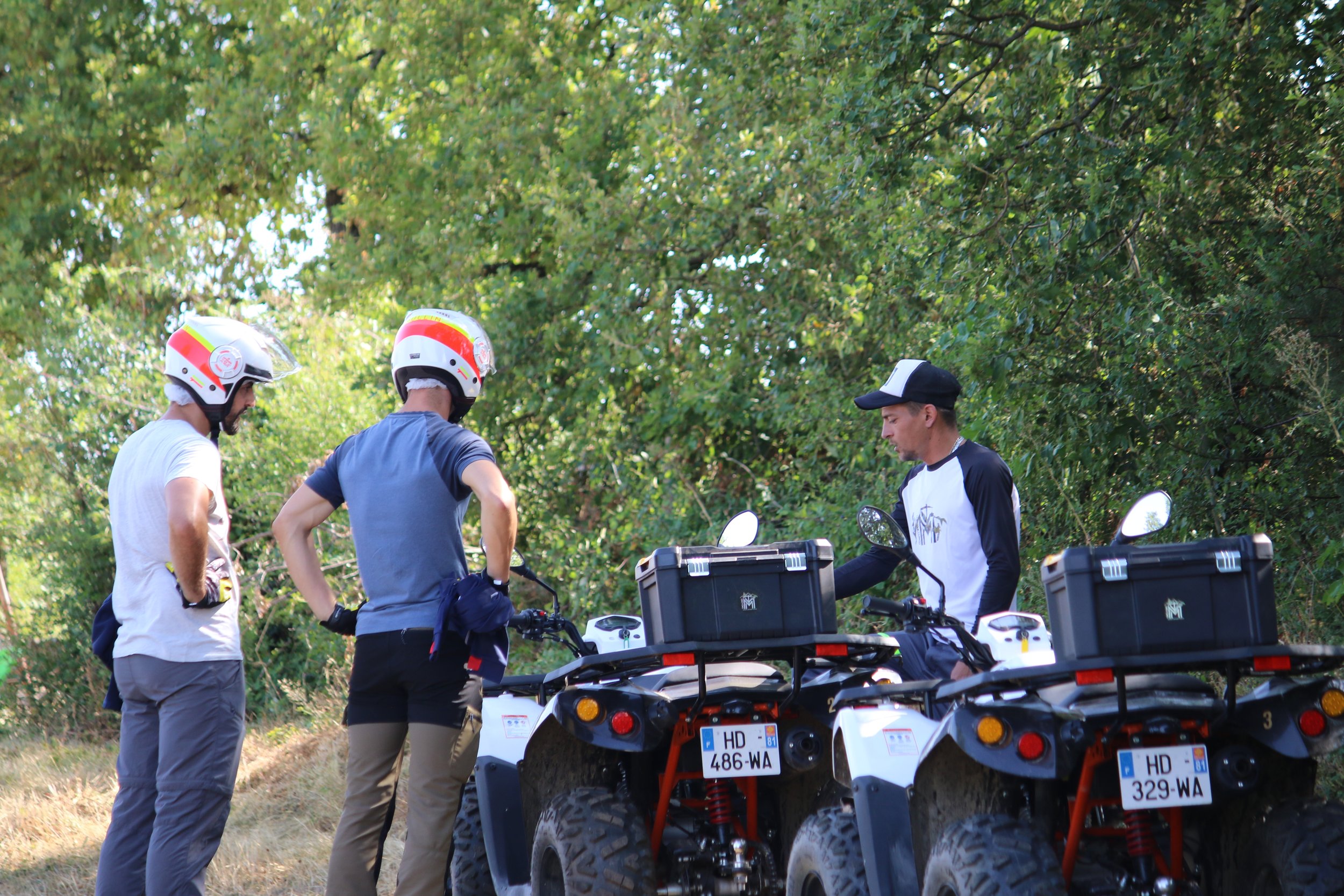 Trois personnes près de deux quads sur un sentier forestier, deux portent un casque et parlent à un homme sans casque avec une casquette. La scène se déroule dans une forêt verdoyante.