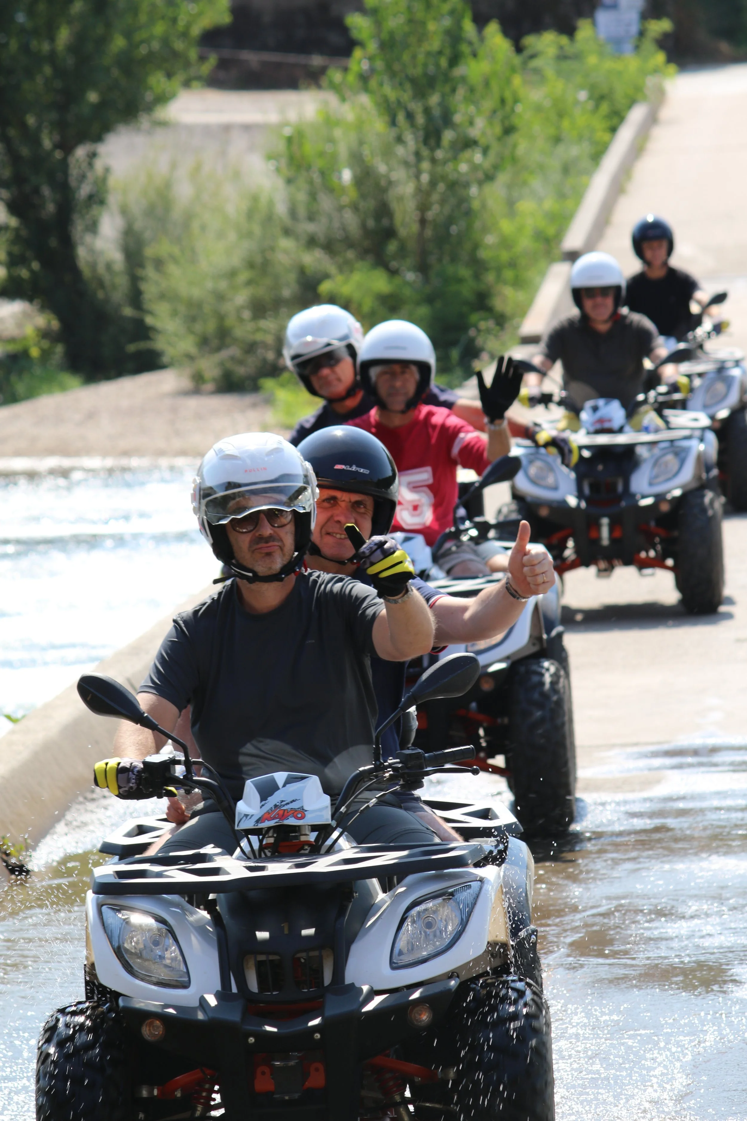 Groupe de personnes en quad, portant des casques, roulant sur un chemin près de l'eau, certains faisant des signes de la main.