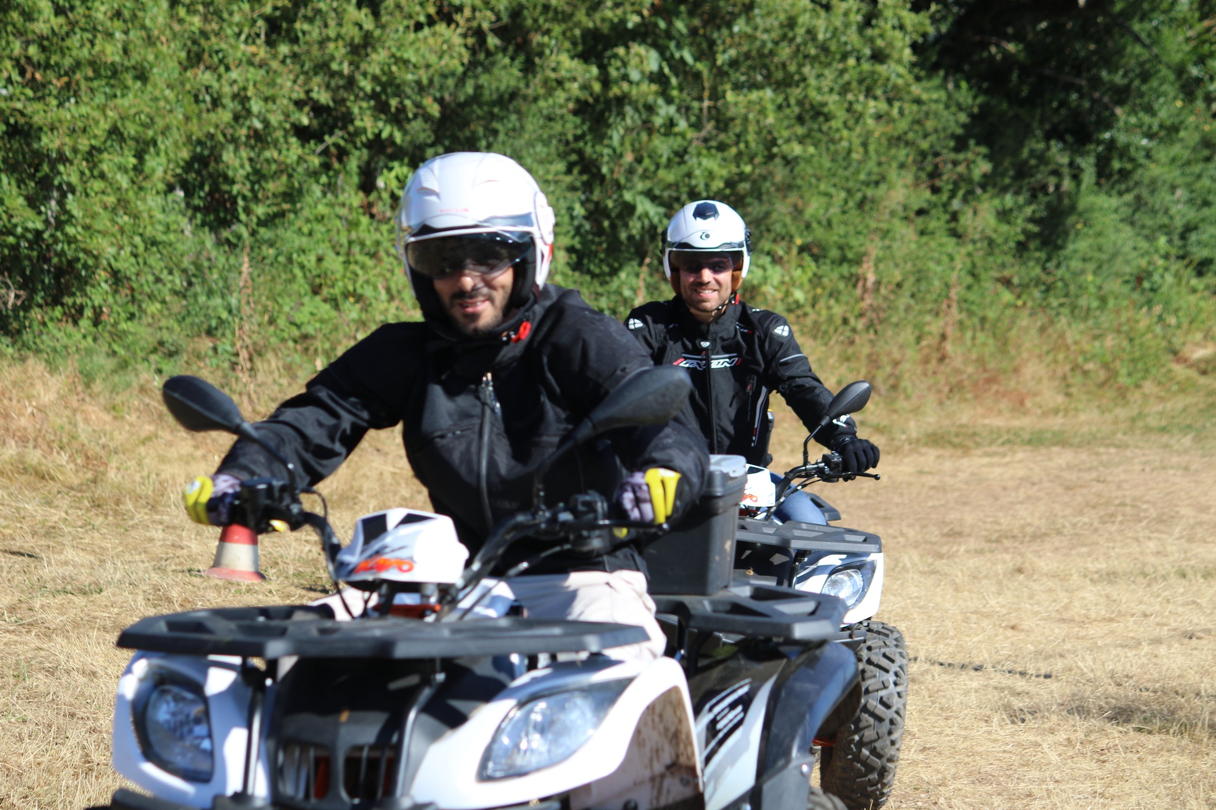 Deux hommes en moto tout-terrain, portant des casques et des vêtements de protection, roulent en groupe dans un champ sec avec des arbres en arrière-plan.