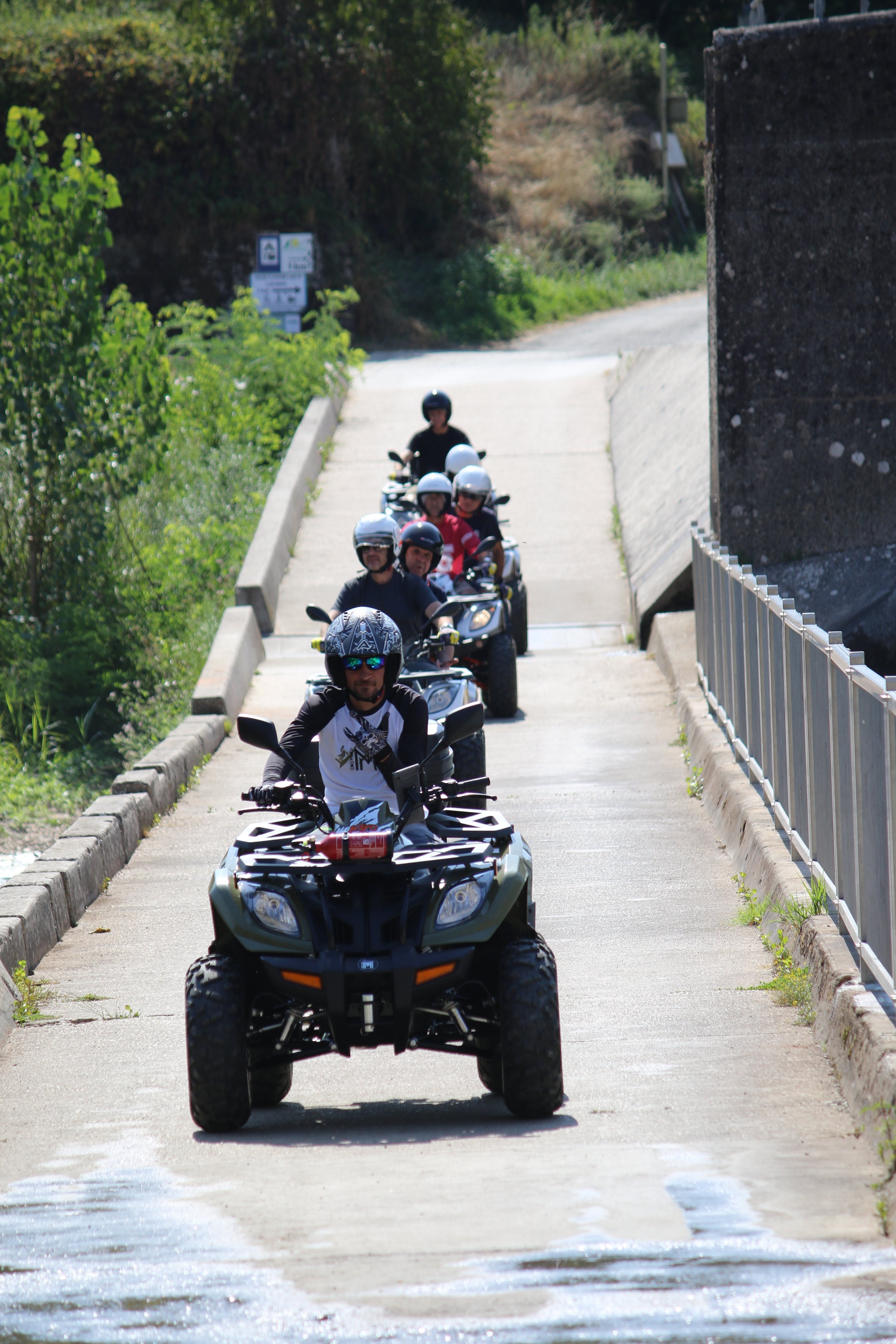 Groupe de personnes à cheval sur des quads, portant des casques, en ligne sur une route escarpée entourée de végétation.