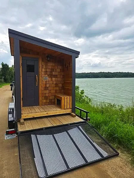 Small wooden sauna on a trailer with a door, bench, and an accessible ramp, located near a body of water with grassy area and cloudy sky.