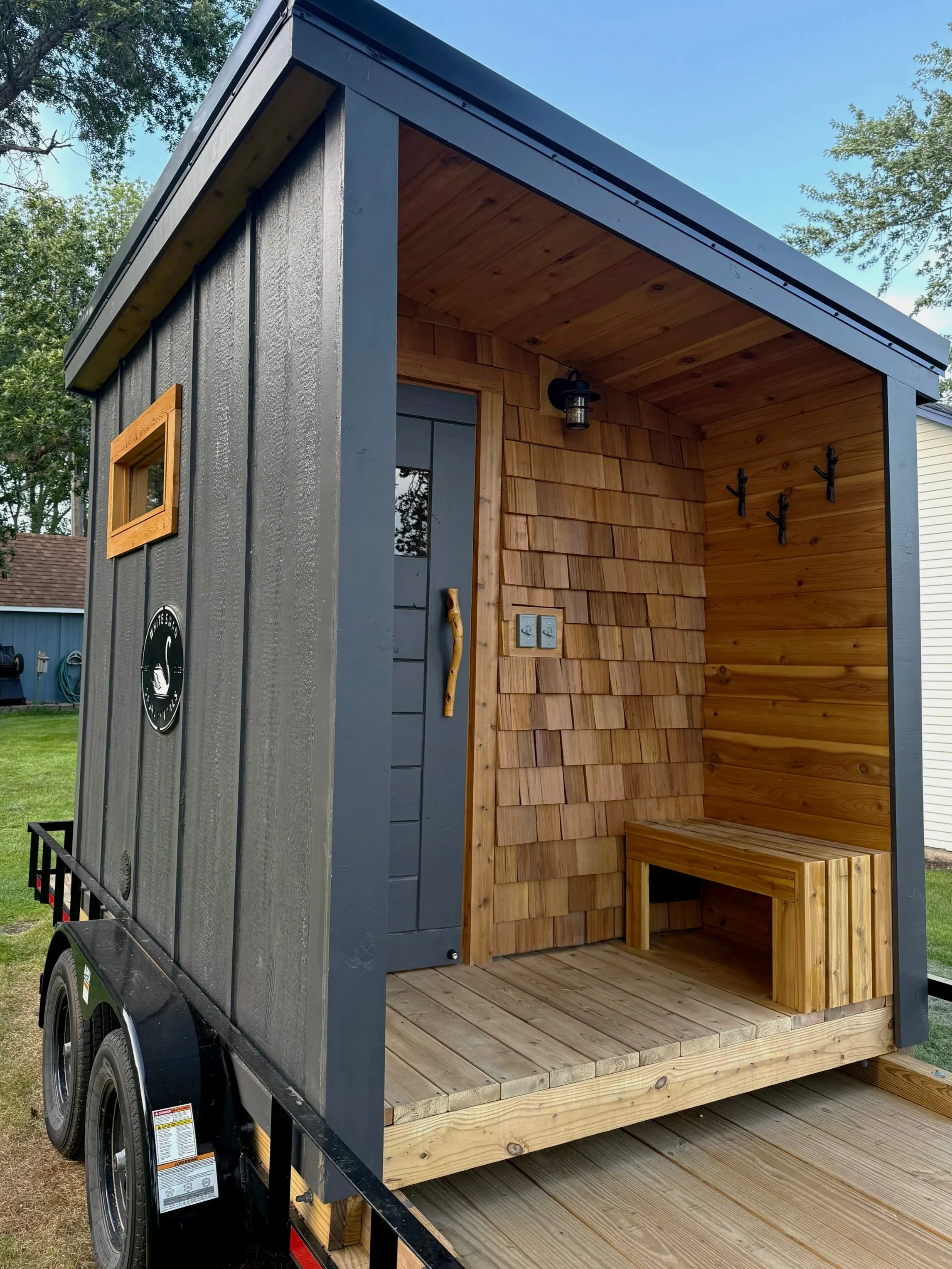 Outdoor sauna with a dark gray exterior, wooden interior, a bench, coat hooks, and an exterior light.