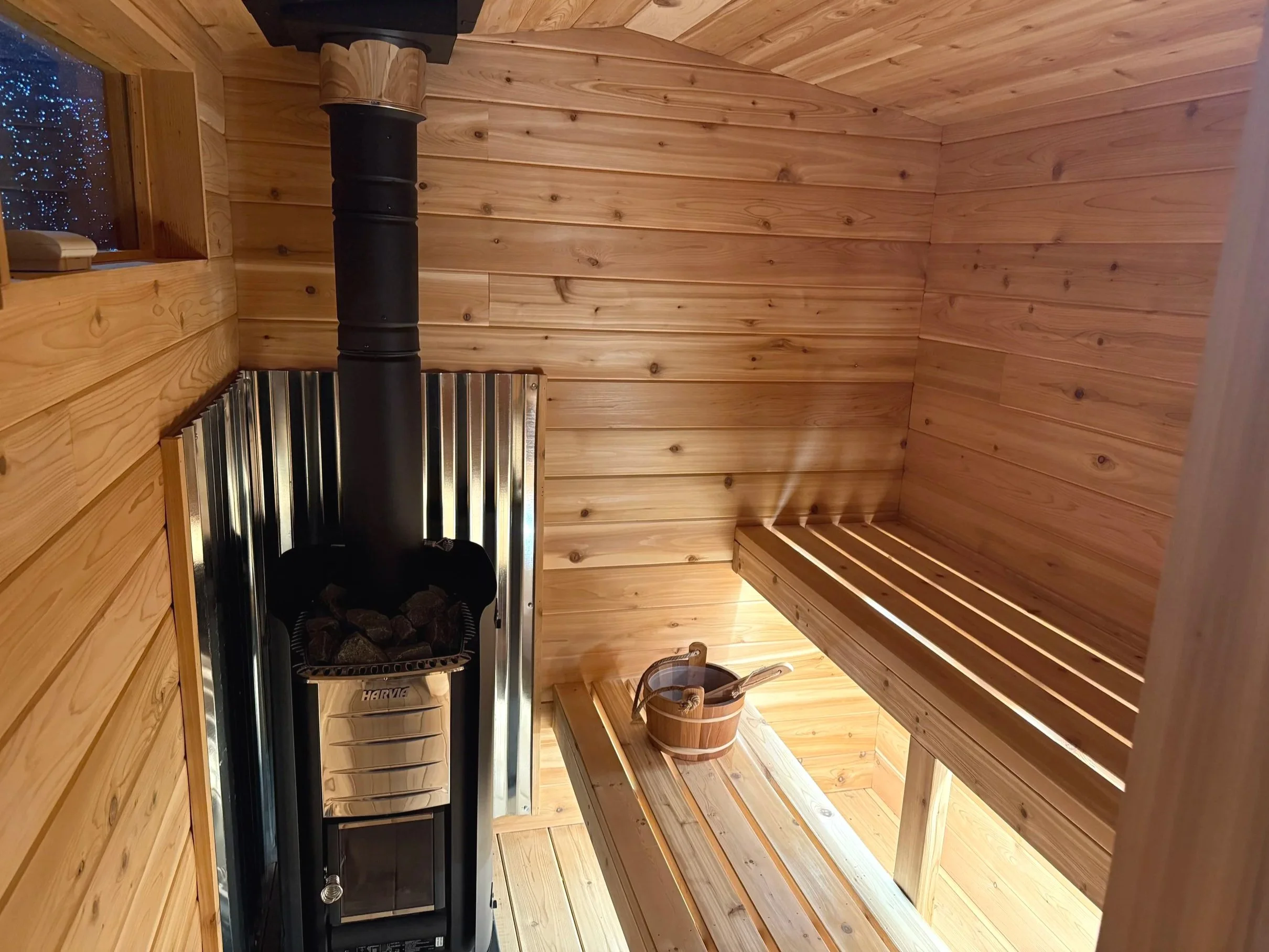Interior of a traditional Finnish sauna with wooden walls, a bench, a wooden bucket with a ladle, and a stove with stones.