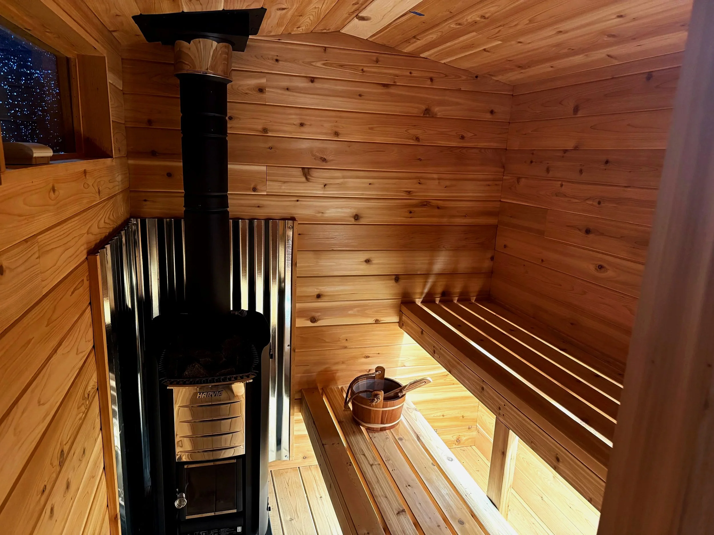 Interior of a wood-paneled sauna with a black stove, a wooden bucket with a ladle, and a bench.
