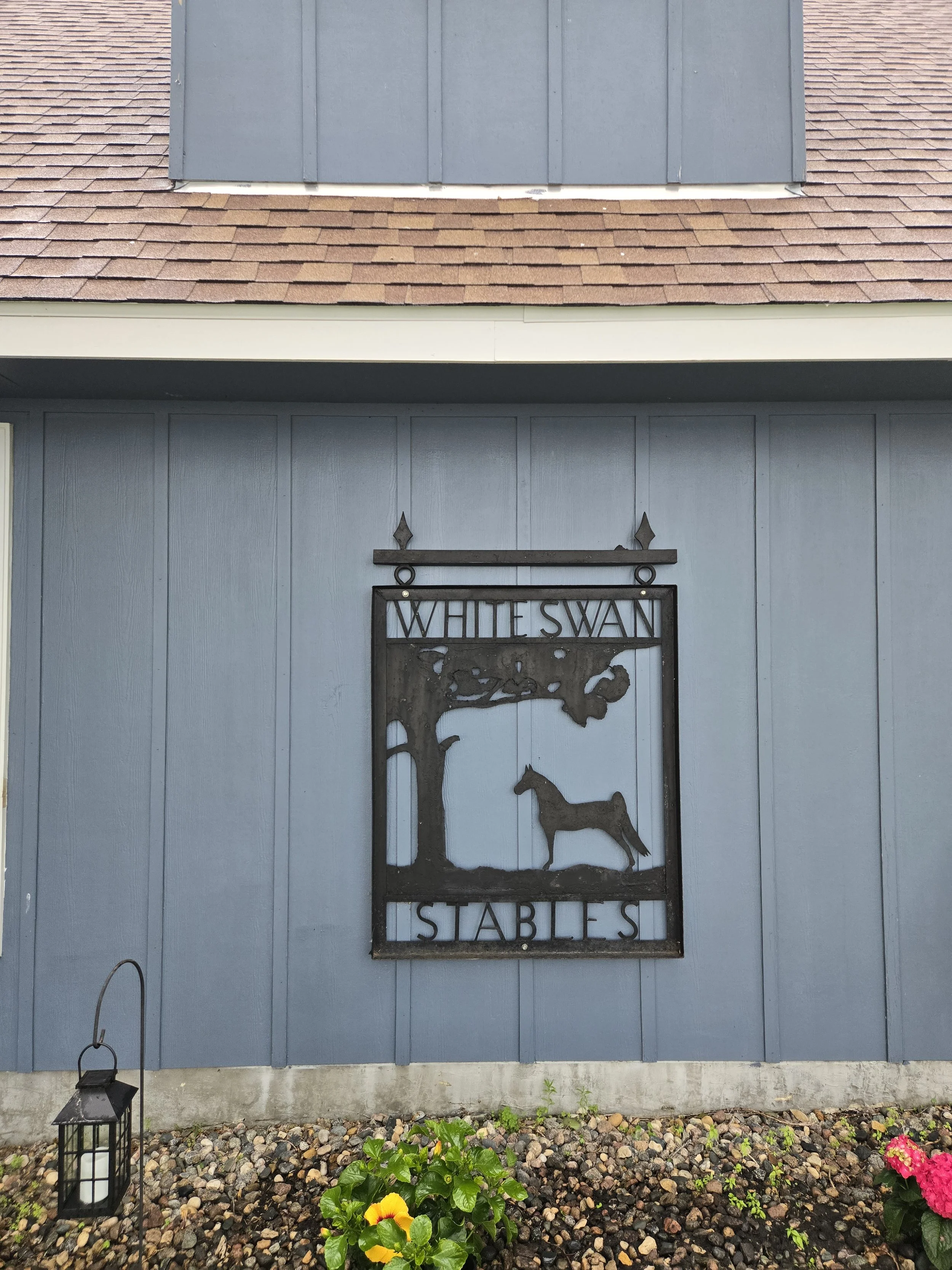 Metal sign for White Swan Stables attached to a light blue wooden barn wall, with a tree and a horse silhouette. A lantern and some colorful flowers are on the ground in front.